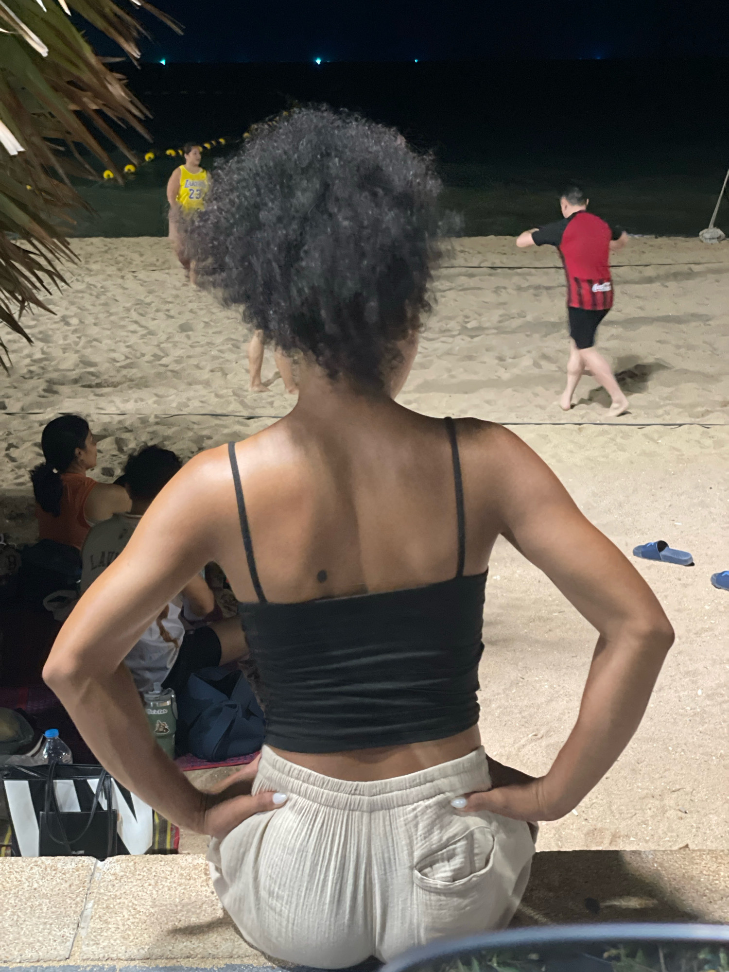 The image depicts a nighttime scene on a sandy beach, likely in Pattaya City, Thailand, with a focus on a woman viewed from behind. She is wearing a black tank top and loose, light-colored pants. Her hair is dark and curly, and she has her hands on her hips. In the background, two men are engaged in a game on the sand court, possibly beach volleyball, with a net visible. One man wears a red and black jersey, while another, further in the background, is wearing a yellow tank top with the number 23. Several people are seated on the sand in the mid-ground. The lighting suggests it's evening or night, with artificial lights illuminating the scene and the ocean in the distance. Palm fronds frame the left side of the image.