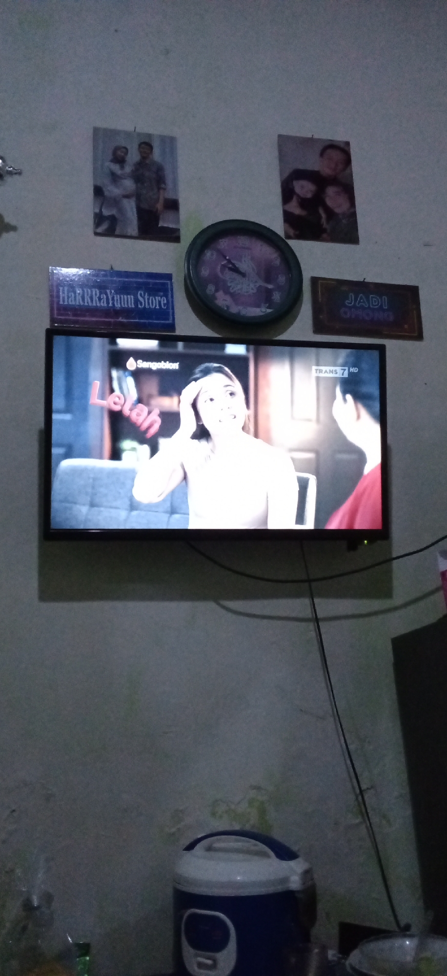 The image shows an indoor scene, likely a living room, with a television displaying a program. The wall behind the television is decorated with several items.
In the upper left of the wall, there are two framed photographs. The left photograph appears to be of a couple, with the woman in profile and potentially pregnant. The right photograph shows a group of three people, likely a family, with two adults and a child.
To the left of the television, there is a rectangular sign with the text "HaRRRaYuuu Store" in blue letters on a blue background. Above the television, slightly to the right, is a round clock with a purple face. To the right of the clock, there is a rectangular framed sign with the text "JADI OMONG" in white letters with a darker background.
The television itself is displaying a program with a woman in the foreground, looking distressed and touching her forehead. Text on the screen reads "Sangobion" and "Lelah" in large pink letters, and "TRANS 7 HD" in the upper right corner. The setting appears to be indoors, with a sofa visible behind the woman.
In the lower portion of the image, a blue and white rice cooker is visible on a surface. A power cord extends from the television and drapes down the wall. The wall itself appears to be a textured, light-colored surface. The overall lighting suggests it is evening or the room is dimly lit.
The provided information indicates the location is Banjaran, Indonesia, but this is not visually discernible from the image itself.