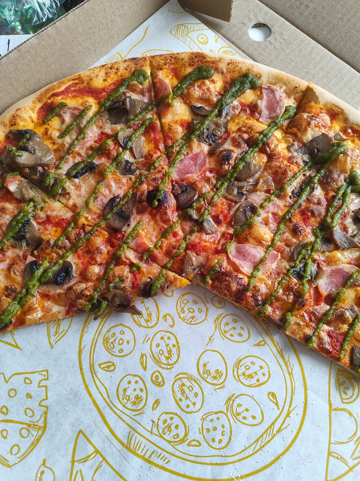 A partially consumed round pizza is observed within an open corrugated cardboard delivery box, located in Marusino, Russia. The pizza, cut into rectangular slices, features a golden-brown crust with some areas of charring. Visible toppings include melted, bubbly cheese, a red tomato sauce base, numerous slices of sautéed mushrooms, and thin, pale pink slices of meat, consistent with ham or bacon. A bright green, viscous sauce, characteristic of pesto, is diagonally drizzled across the entire visible surface of the pizza. The interior base of the cardboard box is lined with white paper adorned with yellow line drawings of pizza slices, whole pizzas, and cheese forms. Small, scattered white particulate matter is present on this paper lining. A brown cardboard lid is partially visible, folded back at the top right, revealing a circular cutout. In the upper left background, a clear plastic bag containing indistinct green material is visible out of focus. No individuals are present.