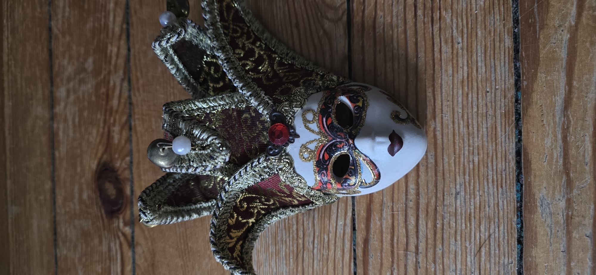 A Venetian-style mask lies diagonally across a wooden surface composed of vertically aligned planks. The mask is predominantly white with decorative patterns around the eye holes in shades of red, black, and gold. A small, painted red heart is visible on the right cheek of the mask. The upper and left side of the mask displays ornate protrusions of red and gold brocade fabric, edged with gold trim. A large red jewel is near the front of one extension. A white pearl attached to a metal pin is visible on the left side. The wooden surface consists of planks with a light brown tone, exhibiting natural grain patterns and slight variations in color. Varna, Bulgaria is not obviously present in this image.