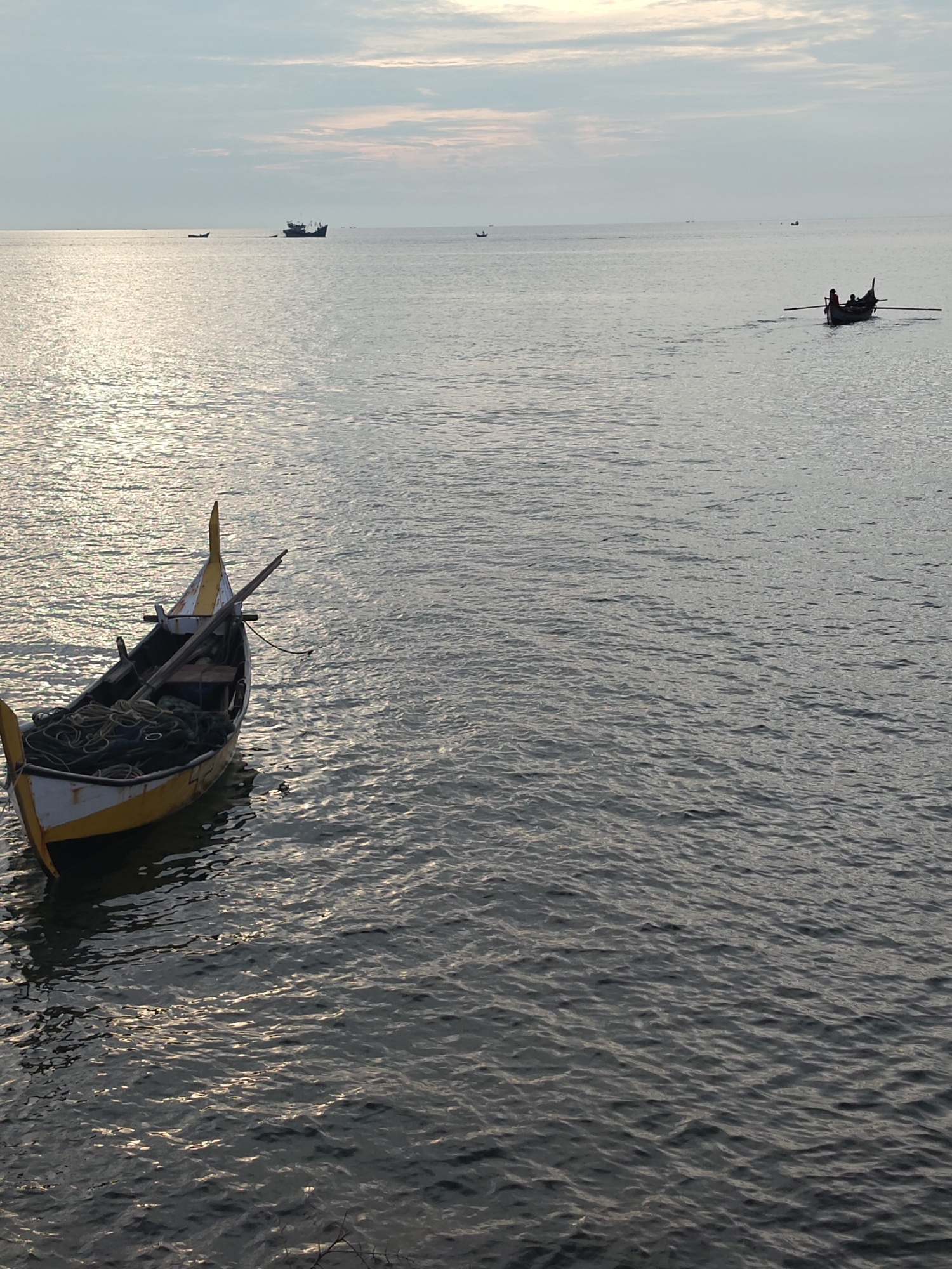 The image depicts a serene ocean scene at what appears to be sunrise or sunset, with the sun's rays reflecting off the rippling water. In the foreground, a traditional fishing boat, painted yellow and white, is anchored and contains coiled ropes and fishing nets. In the distance, another small boat with two occupants rowing is visible. Further out, larger ships can be seen on the horizon. The sky is partly cloudy with soft hues of pink and blue. The overall atmosphere is tranquil, characteristic of the coastal areas around Lhokseumawe, Indonesia, a city known for its fishing industry and proximity to the sea.