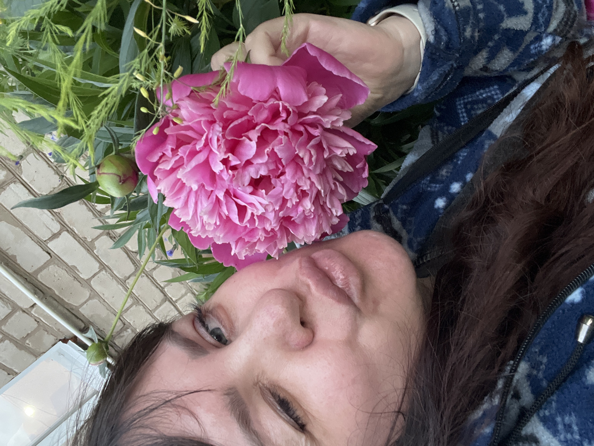 In Panteleevka, Russia, a person with dark hair, wearing a blue and grey patterned jacket, is holding a large, vibrant pink peony flower. The person is looking towards the camera with one eye closed in a wink, and their lips are pursed in a kissing gesture. Their right hand is partially visible, holding the stem of the peony. The background consists of a light-colored brick wall and lush green foliage. A single green peony bud is visible on a stem above and to the left of the person's head. The scene appears to be outdoors.