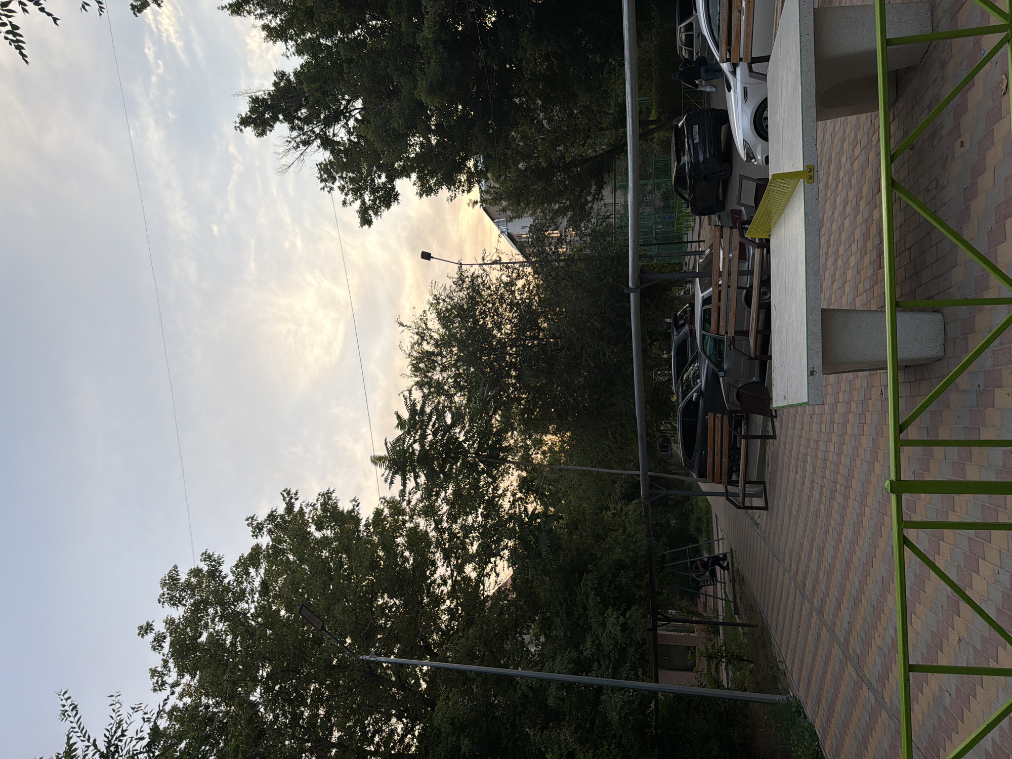 The image depicts a residential courtyard in Shymkent, Kazakhstan. The scene is set during the day, with a cloudy sky transitioning to sunset hues. In the foreground, a concrete ping pong table with a yellow net is visible. Beyond the table, a patterned paved walkway leads to a small playground area with swings. Several parked cars are present in the background, including a white car and a dark SUV. Large trees with dense foliage flank the scene, and a building is partially visible through the trees. A street lamp is positioned between two large trees. The overall atmosphere suggests a typical urban residential setting.