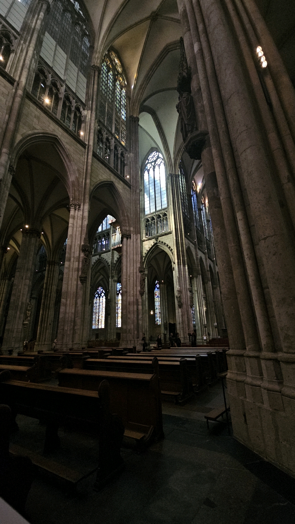 The image depicts the interior of a large Gothic cathedral, likely Cologne Cathedral, in Cologne, Germany. The perspective is low-angle, looking up towards the soaring vaulted ceilings and massive stone columns. Sunlight streams through tall, arched stained-glass windows, casting a colored light onto the stone architecture. Rows of dark wooden pews are visible in the foreground and middle ground, some of them empty, others with a few people seated or standing. In the background, more architectural details of the cathedral's nave and choir are visible, including intricate stonework and more stained-glass windows. The atmosphere is somber and majestic, characteristic of a historic religious building.