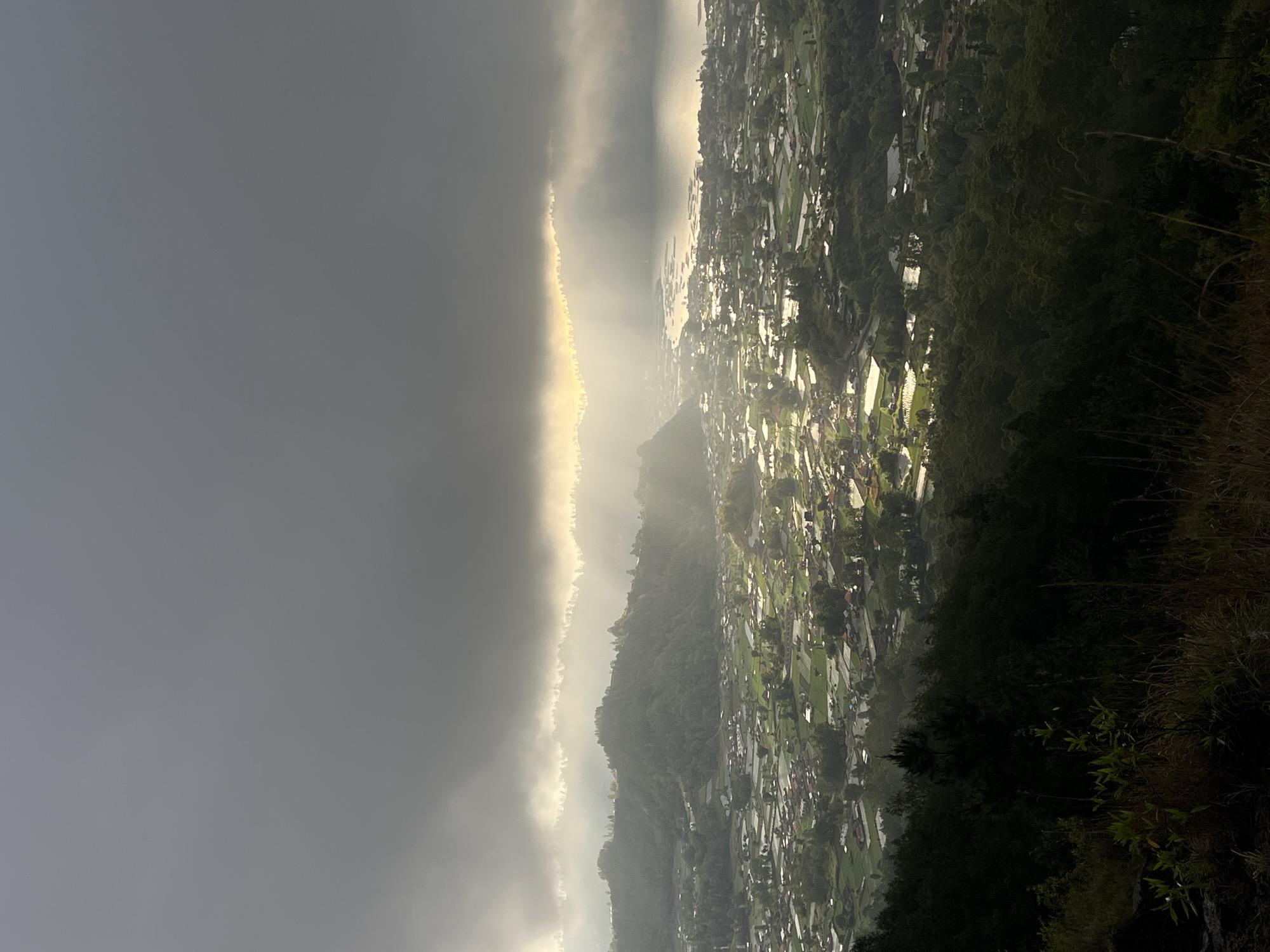 An aerial view captures a valley in Kintamani, Indonesia, with dramatic lighting filtering through dark, ominous clouds. The sun breaks through in the upper center of the frame, casting golden rays down upon the landscape. Below the clouds, a dense green forest covers the foreground hills, transitioning into a sprawling village in the mid-ground. Houses with red roofs are scattered amongst lush green rice paddies, many of which reflect the light. In the distance, a body of water, likely Lake Batur, is visible, with the light of the sun shimmering on its surface. The overall atmosphere is one of a serene, yet dramatic, dawn or dusk in a rural Indonesian setting.