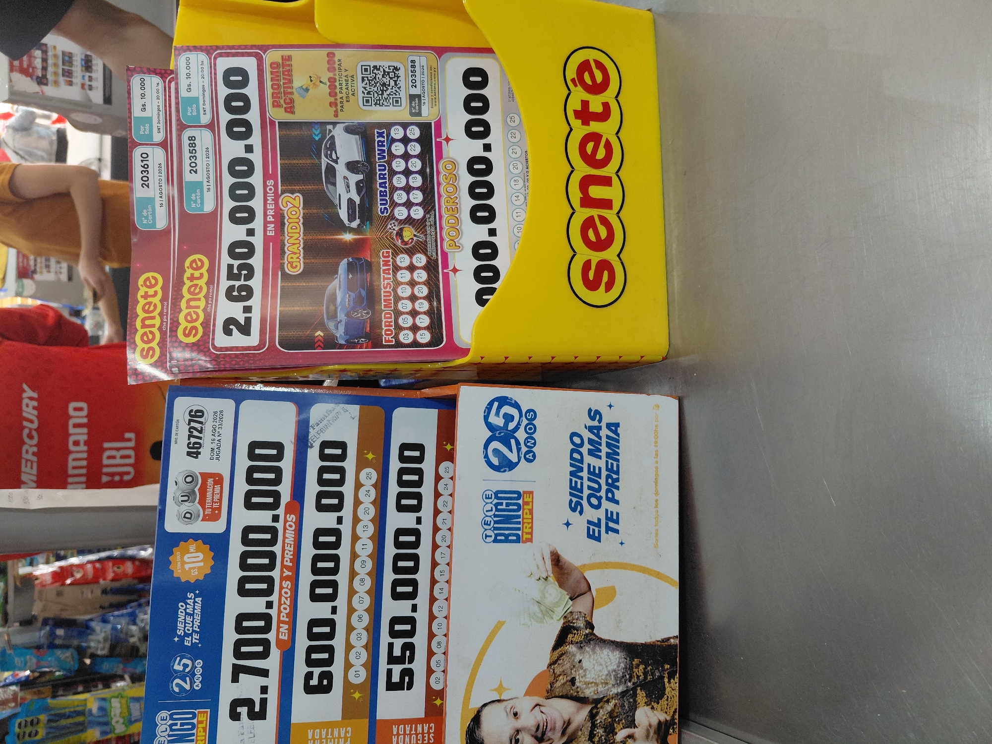 The image shows two lottery ticket displays, likely at a point of sale, possibly in Encarnacion, Paraguay, given the presence of the "Senete" brand and currency notation common in the region. The display on the left is for "Tele Bingo Triple," featuring large prize amounts like 2.700.000.000 and 600.000.000. It highlights "25 Anos" (25 years) and the slogan "Siendo el que más te premia" (Being the one that rewards you the most). The display on the right is for "Senete," with prize amounts such as 2.650.000.000. This display also shows ticket numbers, dates (August 16, 2026), and prices. It features images of cars, including a Ford Mustang and a Subaru WRX, and a QR code for participation. The overall environment appears to be an indoor commercial setting, with a metal counter surface in the foreground. The background shows shelving with various products, including what appear to be electronics and consumer goods.