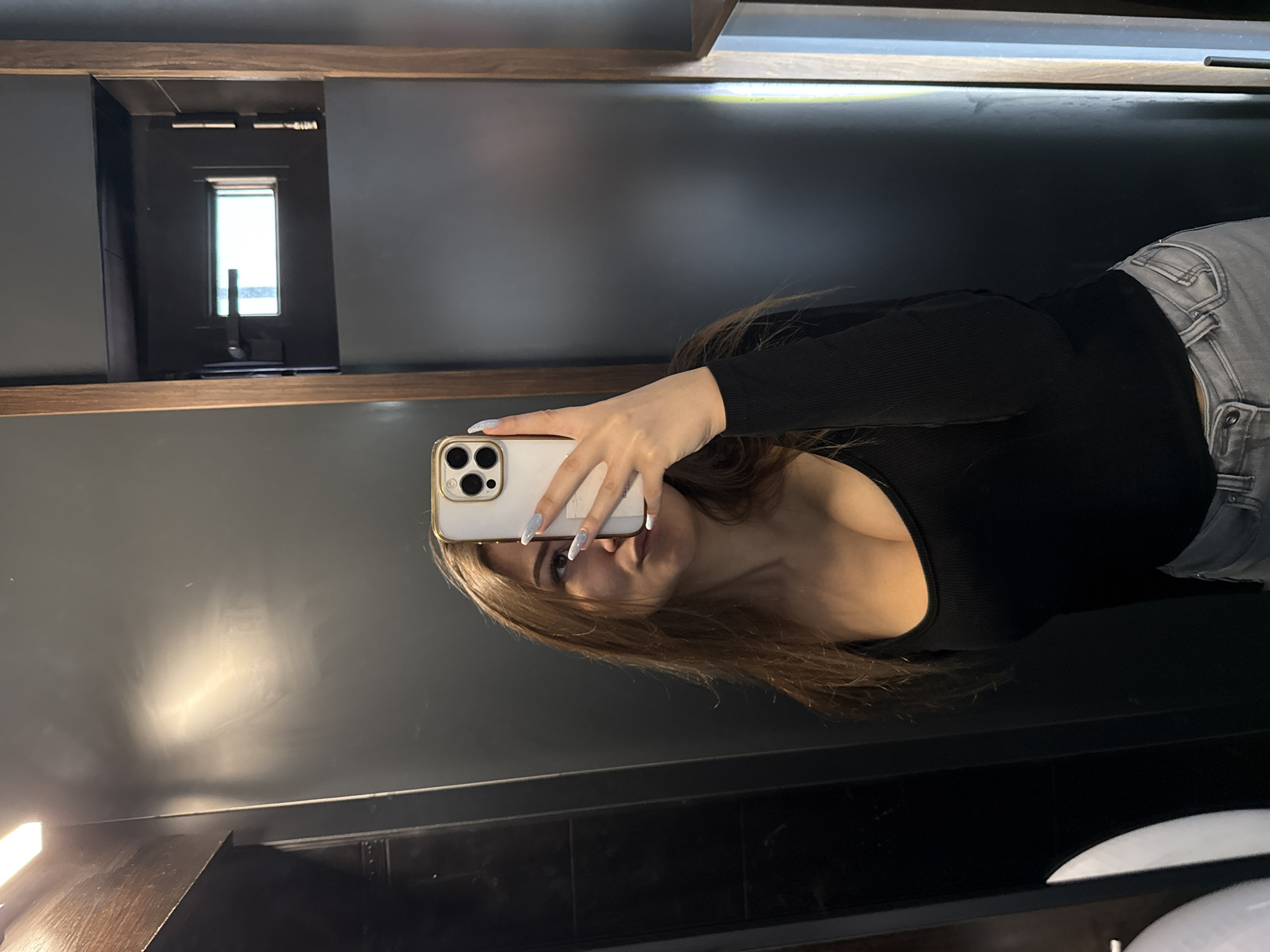 A woman is taking a mirror selfie in a bathroom. The woman has long brown hair, wearing a black top, and gray jeans. She is holding a white and gold iPhone with an elaborate manicure. In the reflection, there is a window and a sink. The bathroom is mostly gray with black accents. The location is not discernible from the image. It is not possible to tell if it is in Sofia, Bulgaria.