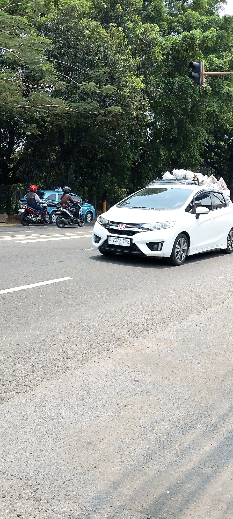 In Depok, Indonesia, a white car is traveling on a road. The car is loaded with several white bags on its roof. In the background, there are trees and a traffic light. Two motorcycles are also visible on the road, with riders wearing helmets. One motorcycle has a rider wearing a red helmet, and the other has a rider wearing a brown helmet. A blue car is parked behind the motorcycles. The road appears to be asphalt, with white lane markings. The scene is under a clear sky.