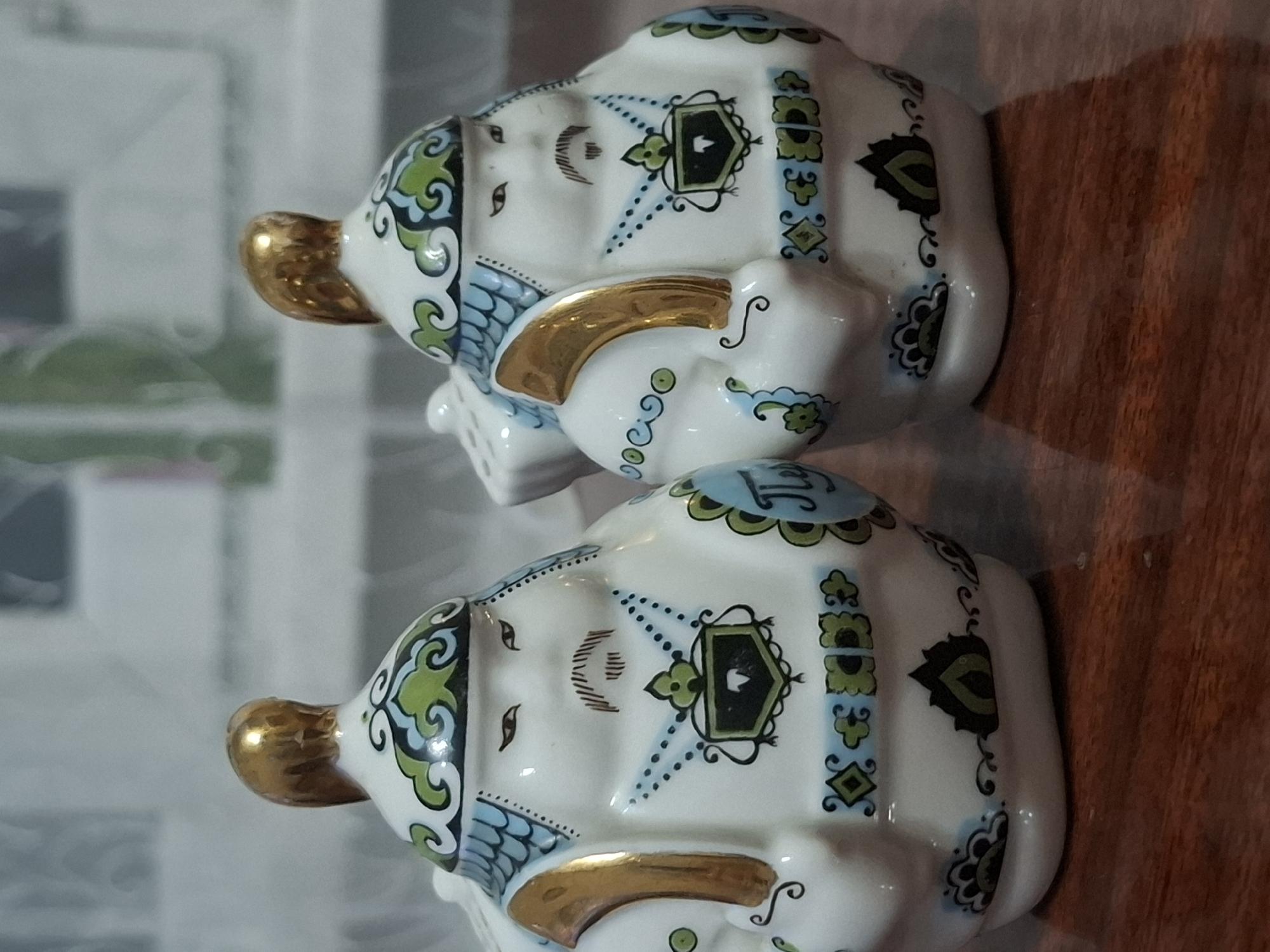 Two ceramic figures, likely salt and pepper shakers, are positioned on a wooden surface. The figures are white with gold accents and painted designs in blue and green, depicting stylized human faces with mustaches and elaborate headdresses. The figure on the left has a gold, sculpted top resembling hair. Both figures have patterned clothing depicted on their bodies, including what appears to be a shield design on the chest of the right figure. The background is blurred, showing what might be a window with a light curtain and greenery. There is no visible indication of the city of Almaty, Kazakhstan, or any specific urban environment. The scene appears to be an indoor still life.
