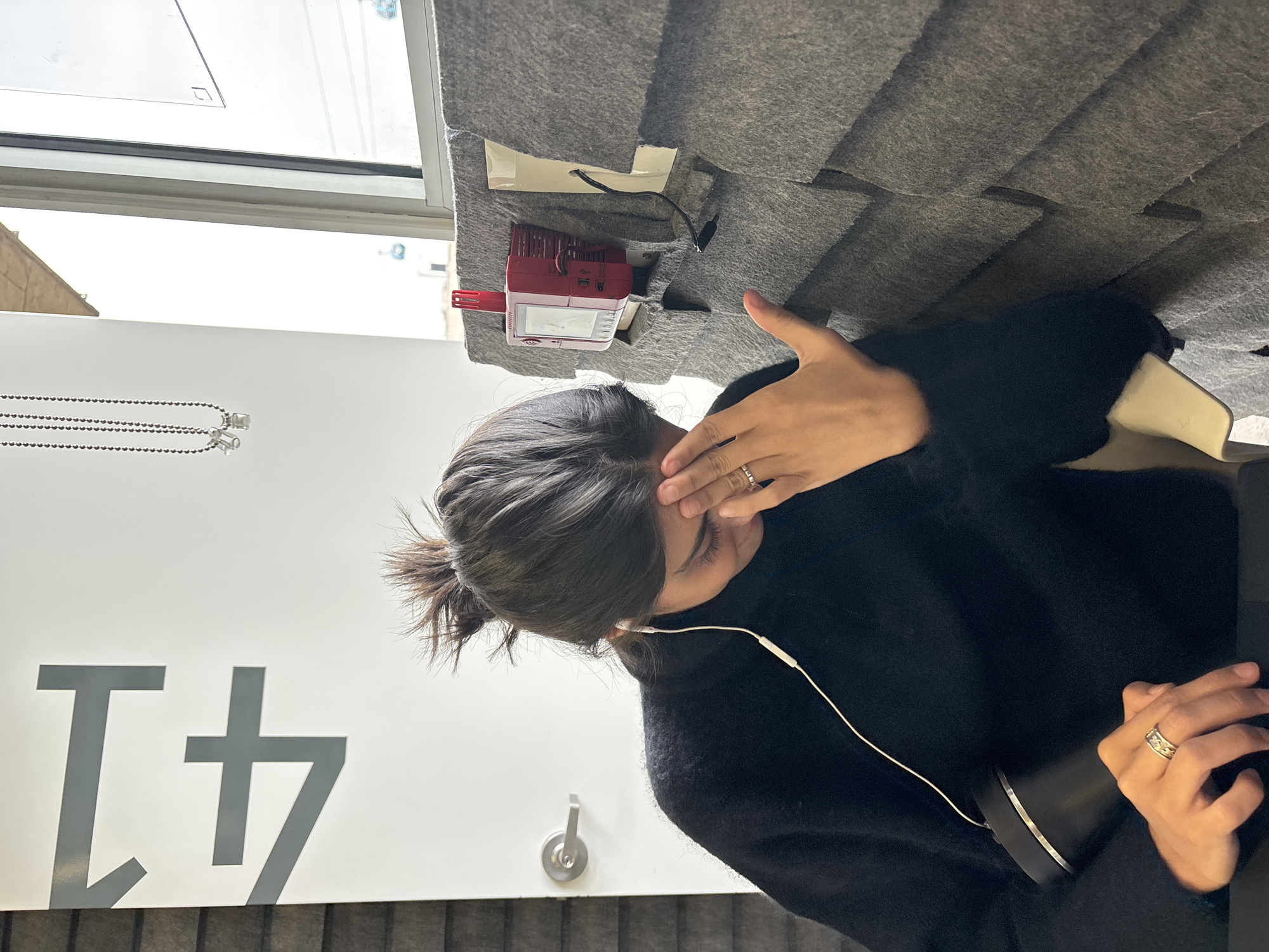 A woman is sitting at a desk with gray acoustic panels. She wears a black sweater and has dark hair pulled back in a ponytail. She is holding her head with one hand and appears to be listening to something through white earphones. On the desk is a red and white device with a screen, and a black cylindrical object, possibly a thermos. A window is in the background. A silver number "41" is visible on the wall to the left of the woman. The scene is located in Mexico City, Mexico.