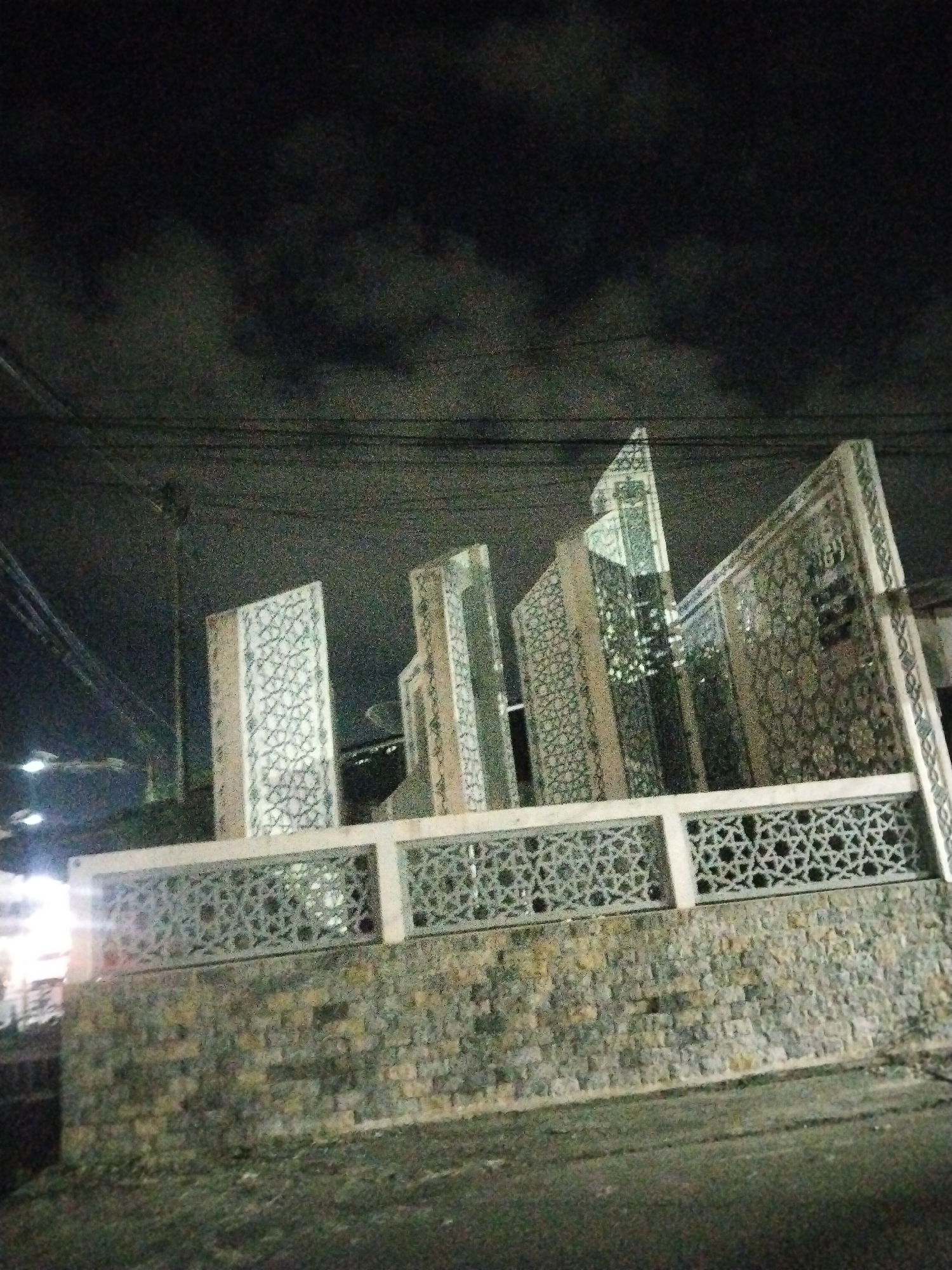The image shows a monument at night in Lhokseumawe, Indonesia. The monument is built on a stone base, topped with a concrete railing featuring geometric cutouts. Above the railing are several vertical panels, also adorned with geometric patterns. Streetlights and power lines are visible in the background, along with dark, cloudy sky. The street in the foreground is dark and appears wet.