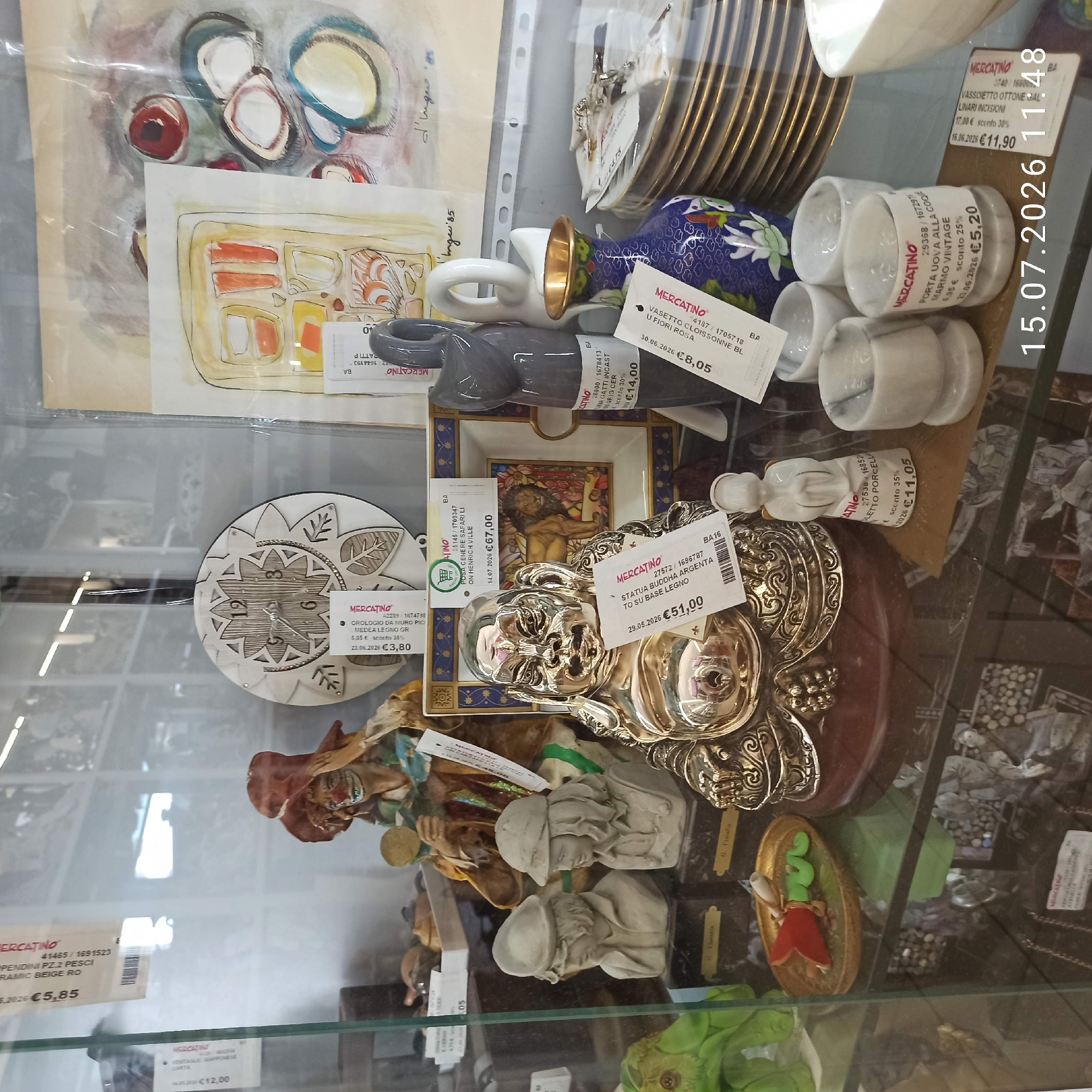 The image shows a display of various items inside a glass-fronted cabinet, likely a store or market stall, in Molfetta, Italy. The items include decorative figurines, plates, artwork, and other miscellaneous objects. In the foreground, a large, ornate golden Buddha statue is prominent, with price tags attached. To the left and right of the Buddha, there are smaller figurines, including busts of two children and a white angel figurine. Behind the Buddha, a decorative plate featuring a lion is visible. In the upper half of the image, abstract paintings and a wooden clock are displayed. Further back, more items are visible, suggesting a cluttered but curated collection. The lighting suggests an indoor environment, possibly a second-hand shop or antique store.