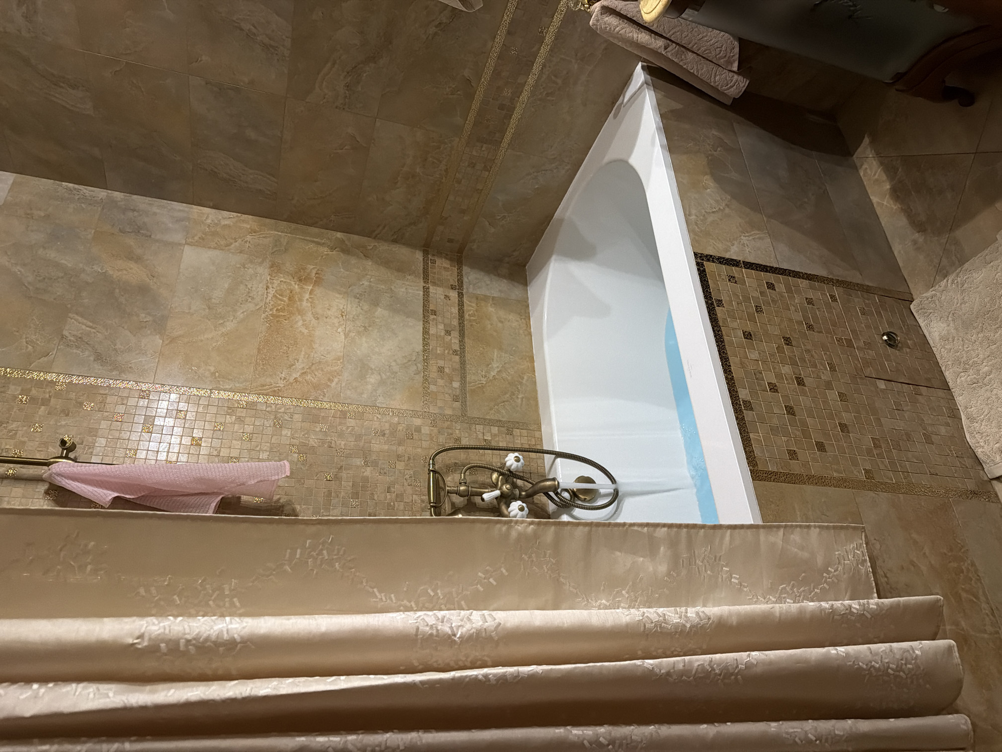 A low-angle, medium shot shows a tiled bathroom with a bathtub set against the right wall. The tiles are primarily in earth tones, with variations in texture and pattern. The bathtub is encased with a decorative fabric overlay that is light beige with a subtle floral pattern. The side of the bathtub is white and extends upwards. The wall behind the tub is covered with smaller square tiles in a brown and beige mosaic. A vintage-style faucet is mounted on the tub, featuring a curved spout and white handles. To the left of the faucet, a towel rack holds a folded pink towel. The ceiling features a similar tile pattern as the walls, with a decorative border running along the center. The foreground consists of the layered fabric adorning the tub. The overall impression is that of a luxurious and well-maintained bathroom. There is no indication of the city or the location of the city of Moscow, Russia.