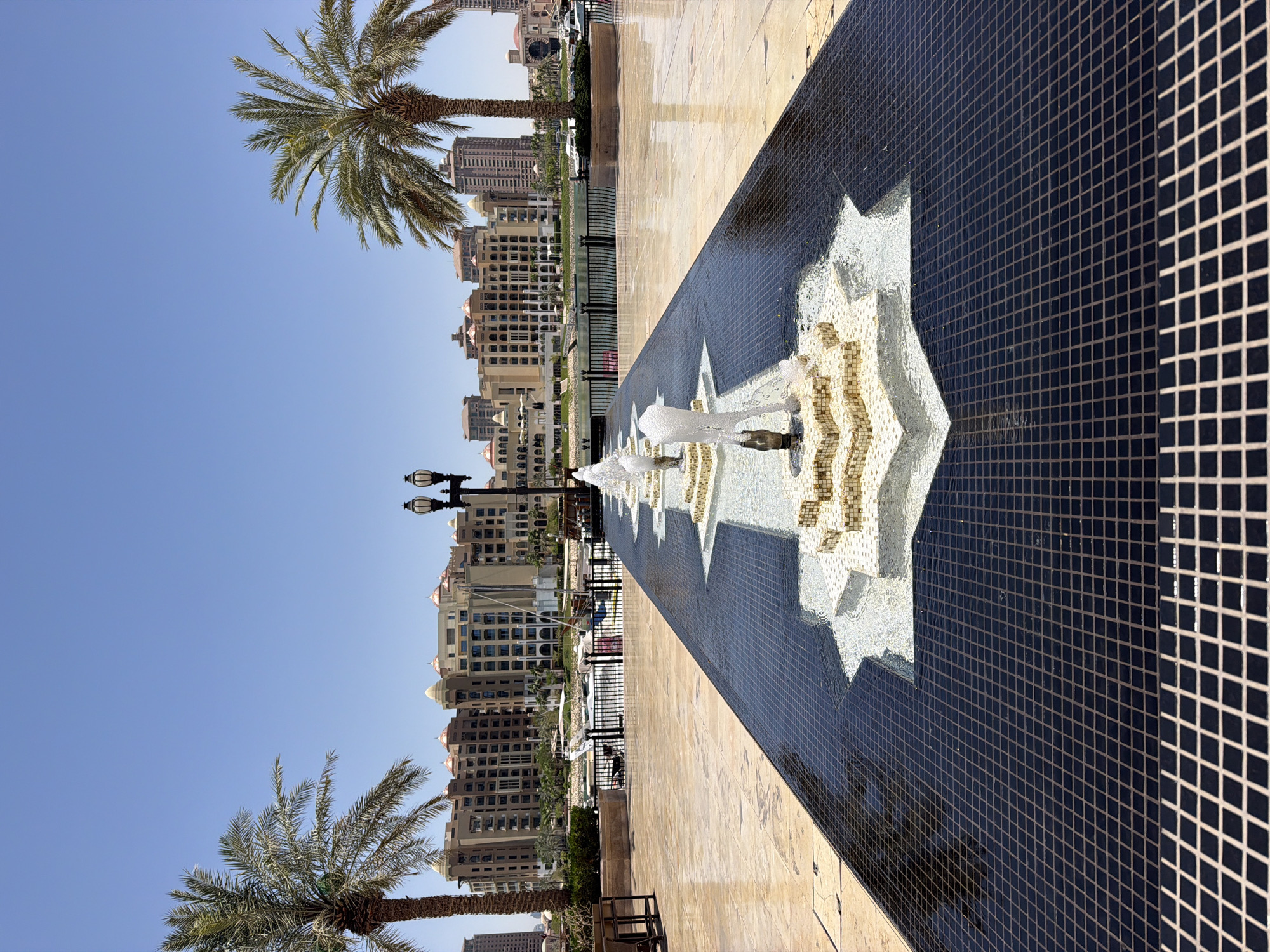 The image shows a long, narrow fountain with water jets in a paved outdoor area. The fountain is lined with black tiles and has decorative star-shaped elements made of light-colored tiles. In the background, there are palm trees, a body of water with boats, and a row of large, tan buildings with arched windows and domes under a clear blue sky. A lamppost stands between the palm trees on the left side of the fountain. The ground is paved with large, light-colored tiles. The scene appears to be in a waterfront area of a city.