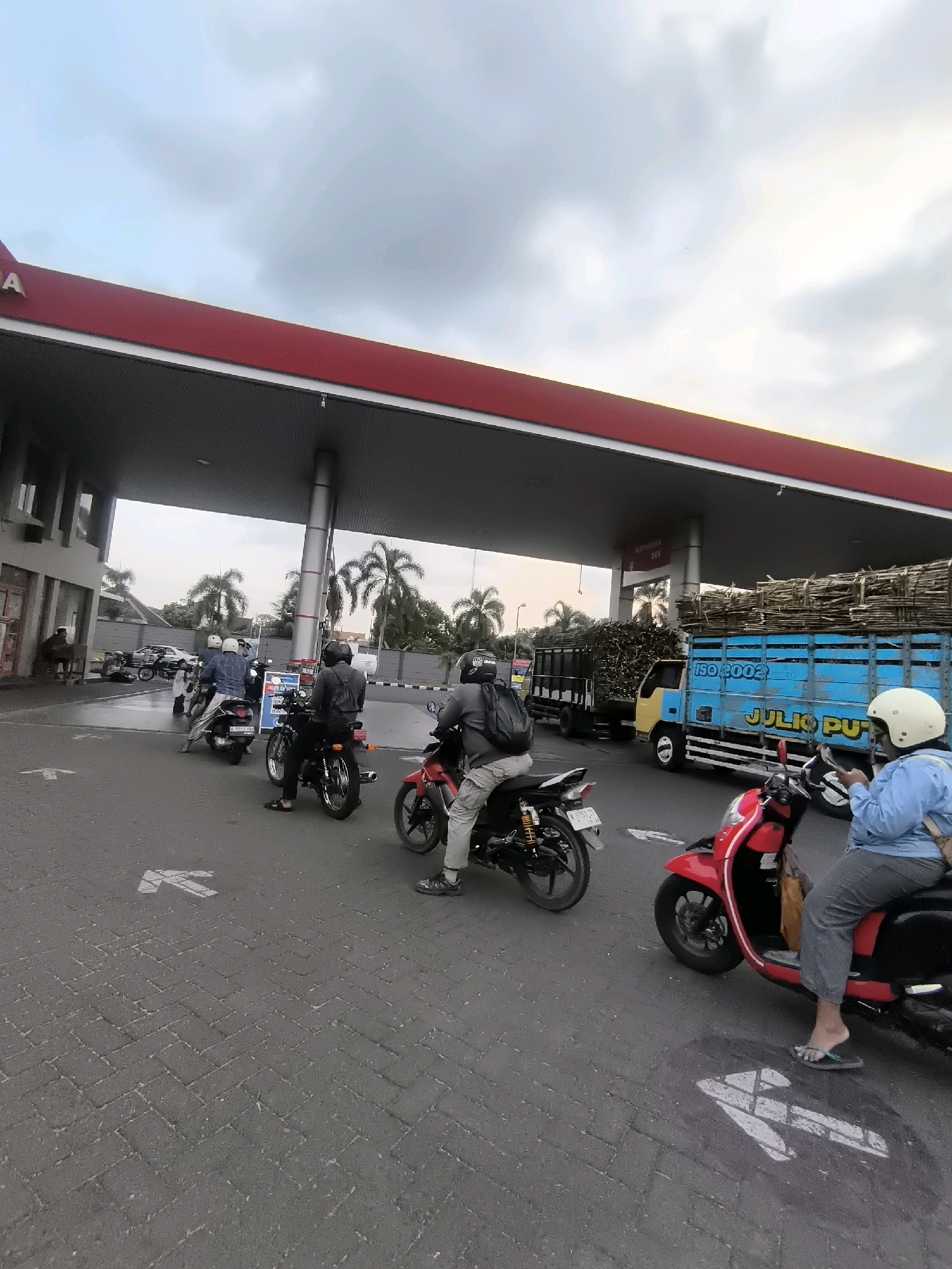 The image depicts a gas station in Kutorenon, Indonesia, under a cloudy sky. A red and white canopy covers several fuel pumps. A blue truck loaded with bundles of wood is parked to the right. In front of the truck, a red scooter with a person wearing a blue jacket and a white helmet is stopped, facing left. To the left of the scooter, two motorcycles are lined up, with riders wearing helmets and casual clothing. One motorcycle is black and the other is black and silver. In the background, palm trees are visible. The ground is paved with interlocking stones. White arrows are painted on the pavement, indicating directions.