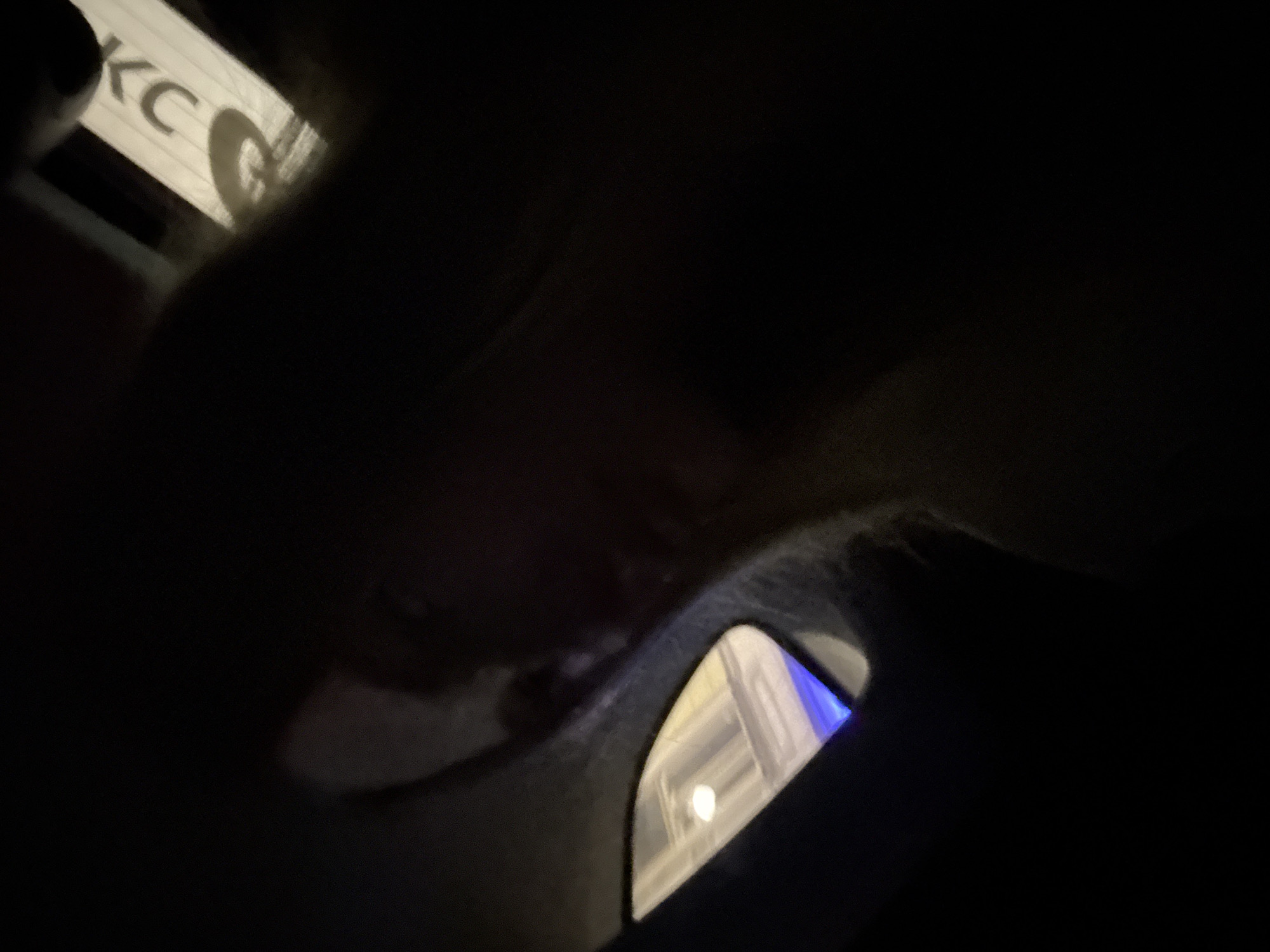 The image shows a dark interior, possibly of a car, with a person's face dimly visible in the center. A small side window reflects a bright, illuminated interior with some blue light at the bottom. A white sign with black lettering, partially obscured, is visible to the upper right. The scene is dark and the details are difficult to discern.
