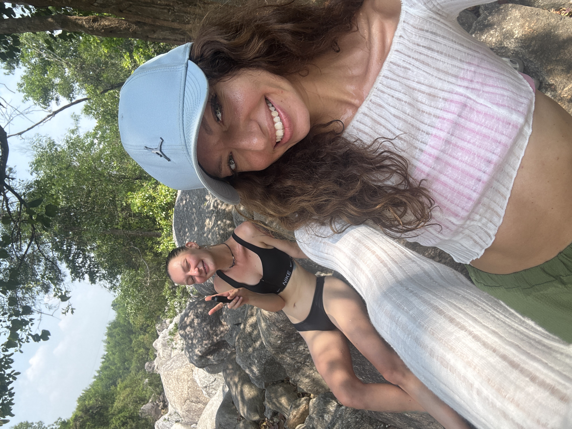 Two women pose for a selfie in an outdoor setting. The woman in the foreground, presumably taking the photo, wears a light blue baseball cap, a white cropped top with pink details, and green shorts. She has brown curly hair and is smiling broadly. Behind her and to the left, the second woman is wearing a black sports bra and bikini bottoms, sitting on rocks. She is making a peace sign with her hand and also smiling. The background includes trees and foliage, suggesting a natural environment, possibly near a beach or river. The photo is taken at an angle, with the camera tilted. It is impossible to determine the location within the city of Đà Nẵng with only this image.
