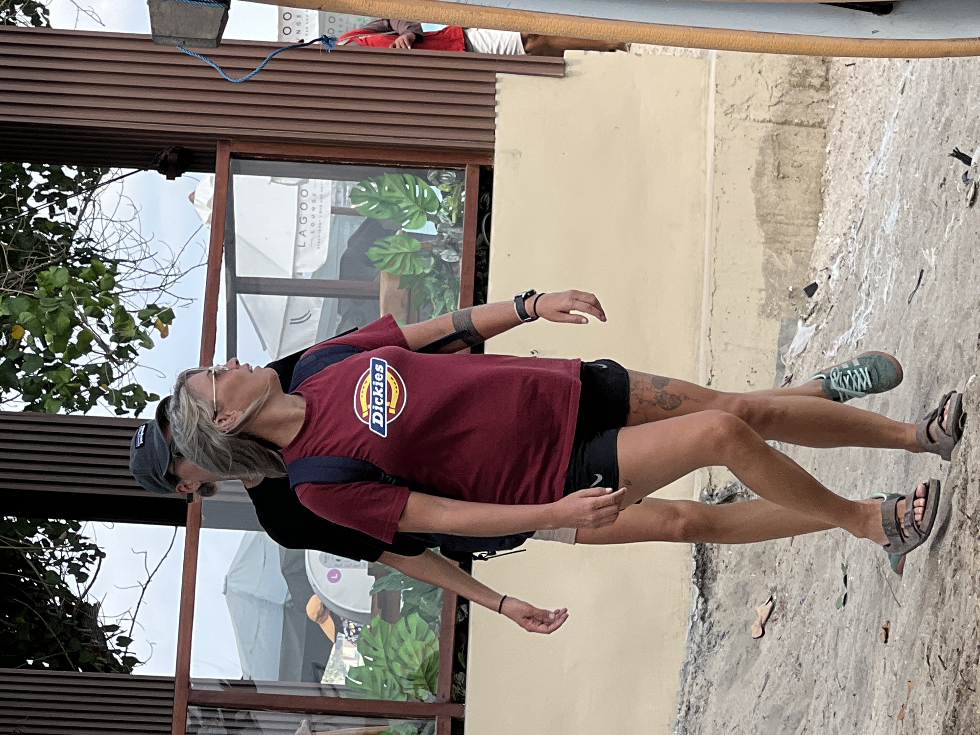 Two individuals are walking on a sandy area near a building with vertical wooden slats and a glass window. The person in the foreground is a woman with blonde hair and glasses, wearing a maroon t-shirt with a Dickies logo, black shorts, and sandals. She has tattoos on her left arm and right leg. She is carrying a navy blue backpack. The person behind her is wearing a dark t-shirt and a grey baseball cap, also with a backpack. The environment appears to be outdoors, possibly in a commercial area. The building in the background has a sign that partially reads "LAGOO LOUNGE BEACHFRONT BAR AND RESTAURANT." The ground is covered in sand and some debris. A person in a red shirt and white shorts is partially visible behind a wooden pole on the right side of the frame.
