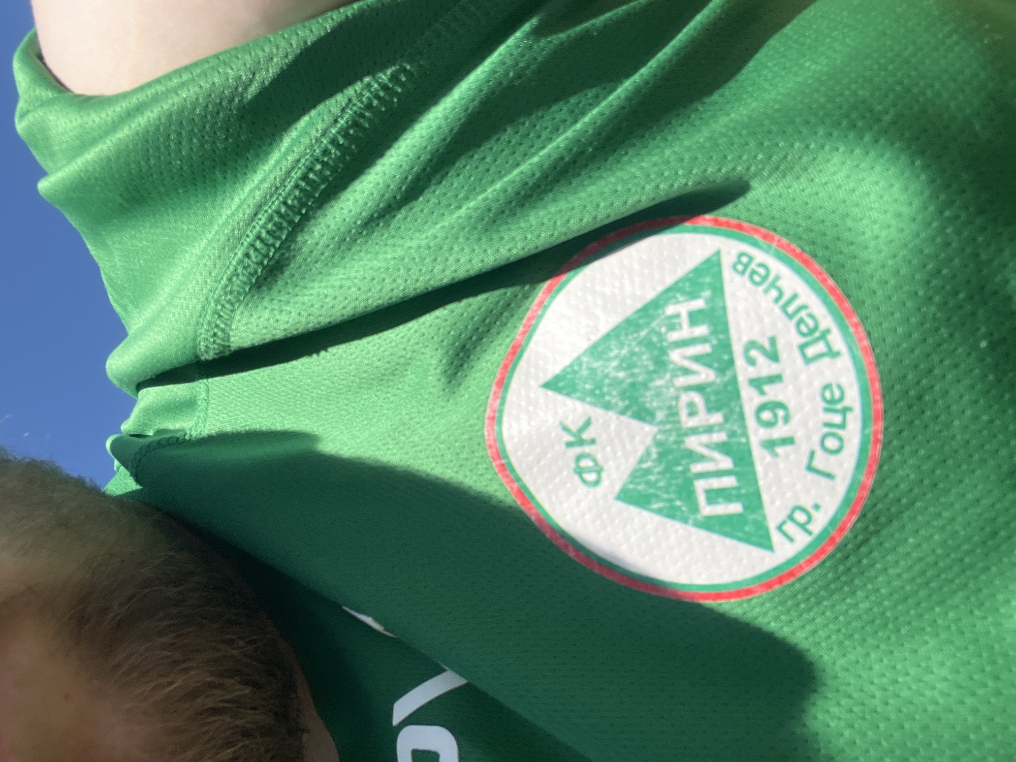 The image shows a close-up view of a green athletic shirt or jersey with a team logo on the right side. The logo is circular, with a white center and a red border. Inside the white area, there is a green triangle pointing upwards. The text "ФК ПИРИН" is arranged on top of the triangle. The text "1912" is below the triangle, and "гр. Гоце Делчев" is at the right. The shirt appears to be made of a mesh-like material. The photo is taken from an angle looking down, revealing a person's hair and the sky in the background. The location can be inferred to be connected to the FK Pirin football club, whose name is featured prominently on the jersey. The shirt represents a team or organization associated with Gotse Delchev, Bulgaria. The image is likely taken in a bright, outdoor setting.
