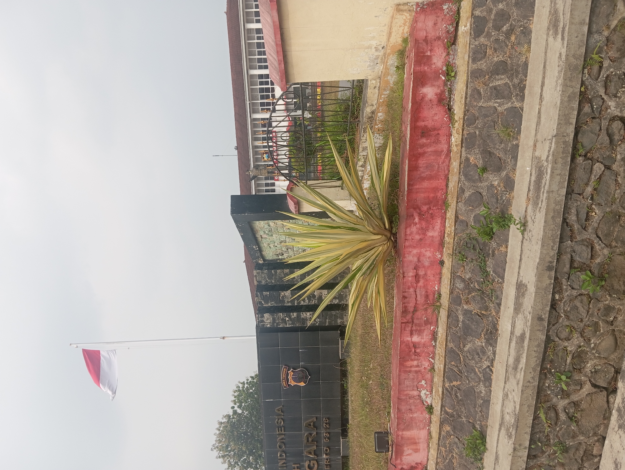 In the city of Sumampir, Indonesia, an outdoor scene features a large, dark monument with white text that reads "INDONESIA". A red and white flag, likely the Indonesian flag, flies from a tall flagpole to the left of the monument. A plant with variegated yellow and green leaves stands in front of the monument. Behind the monument, a building with many windows is visible, and a black metal fence encloses part of the area. The ground is covered in dry grass and a red-painted retaining wall. In the foreground, a stone wall and a concrete structure suggest a drainage system. The sky is overcast and pale.