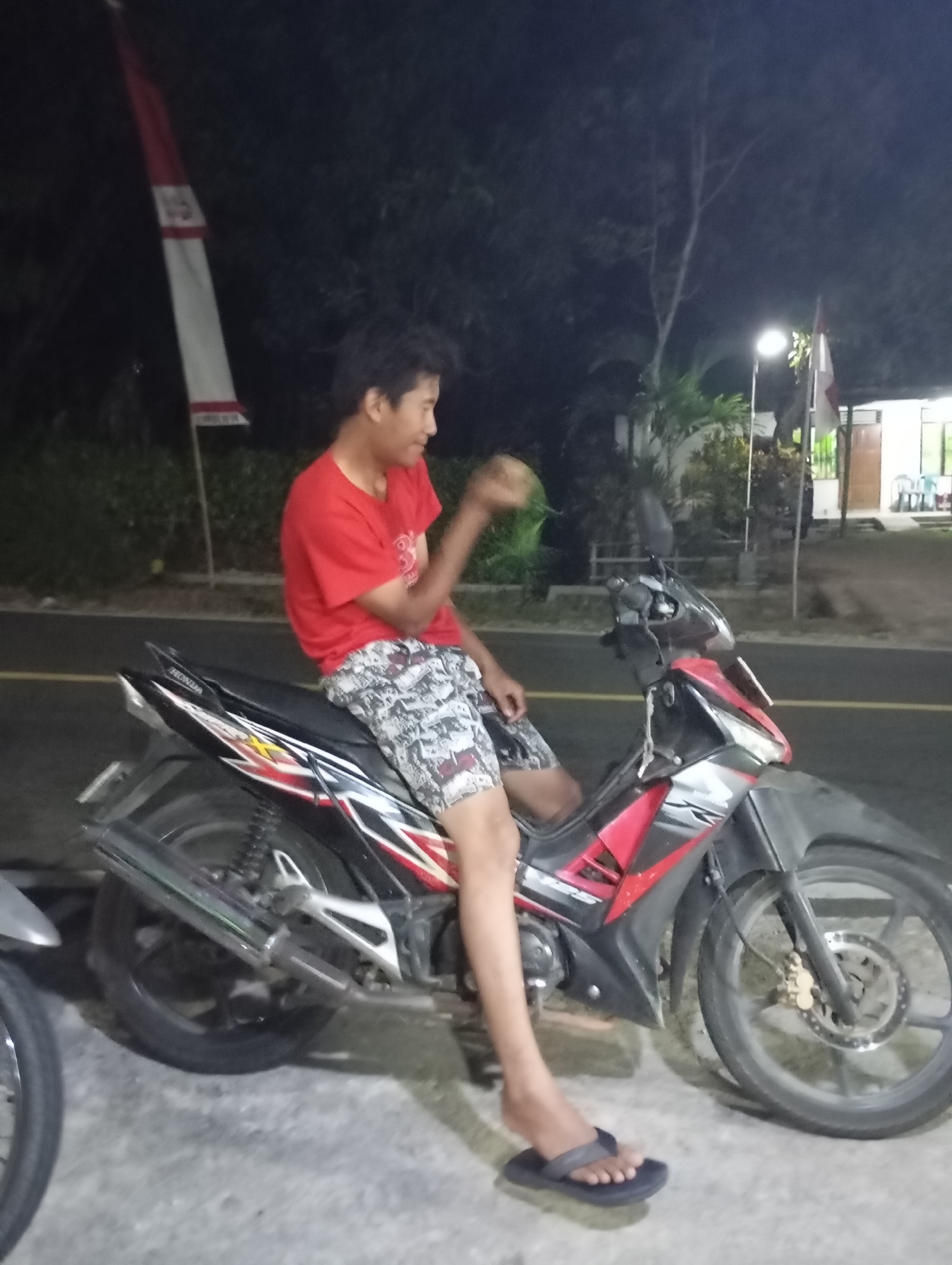 The image shows a young male sitting on a black and red motorcycle at night. He is wearing a red t-shirt and patterned shorts. He is holding a light-colored object in his right hand and appears to be looking to his right. The motorcycle is parked on what appears to be a paved surface, possibly a road or sidewalk, with a yellow line visible on the road. In the background, there is a flag pole with a red and white flag, trees, and a building. The scene suggests an outdoor setting in Wonogondo, Indonesia.