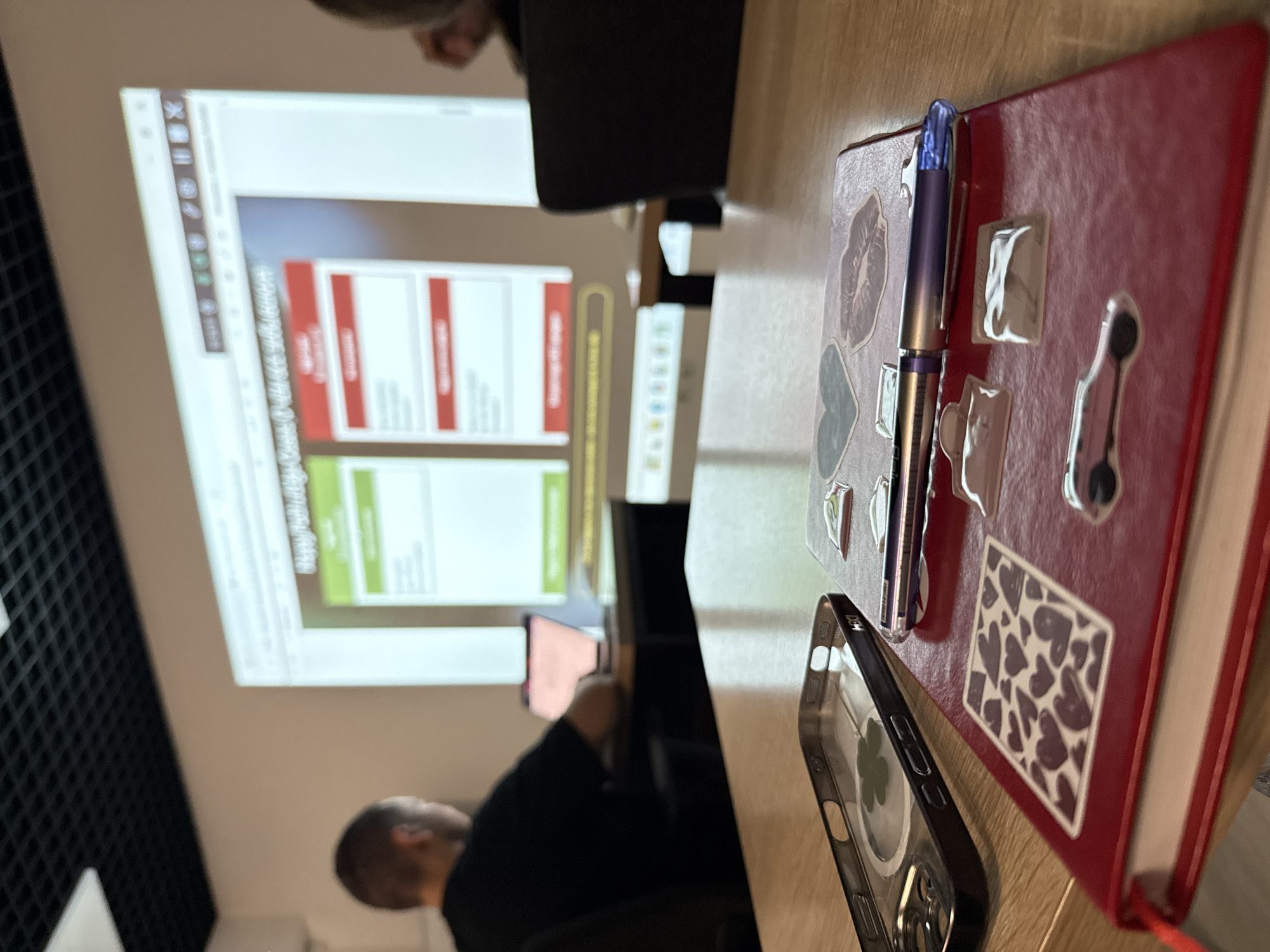 A close-up, eye-level shot shows a red journal with heart stickers and a pen on a wooden table. To the left of the journal is a clear phone case with a four-leaf clover inside. In the background, a projector screen displays a website with red and green sections. A person wearing a black t-shirt is seen in profile, looking at a laptop in front of the screen. The room has a black tiled ceiling and beige walls. The scene is likely an educational or presentation setting, possibly in an office or classroom, but there is no specific information to place it in Bishkek City, Kyrgyzstan.