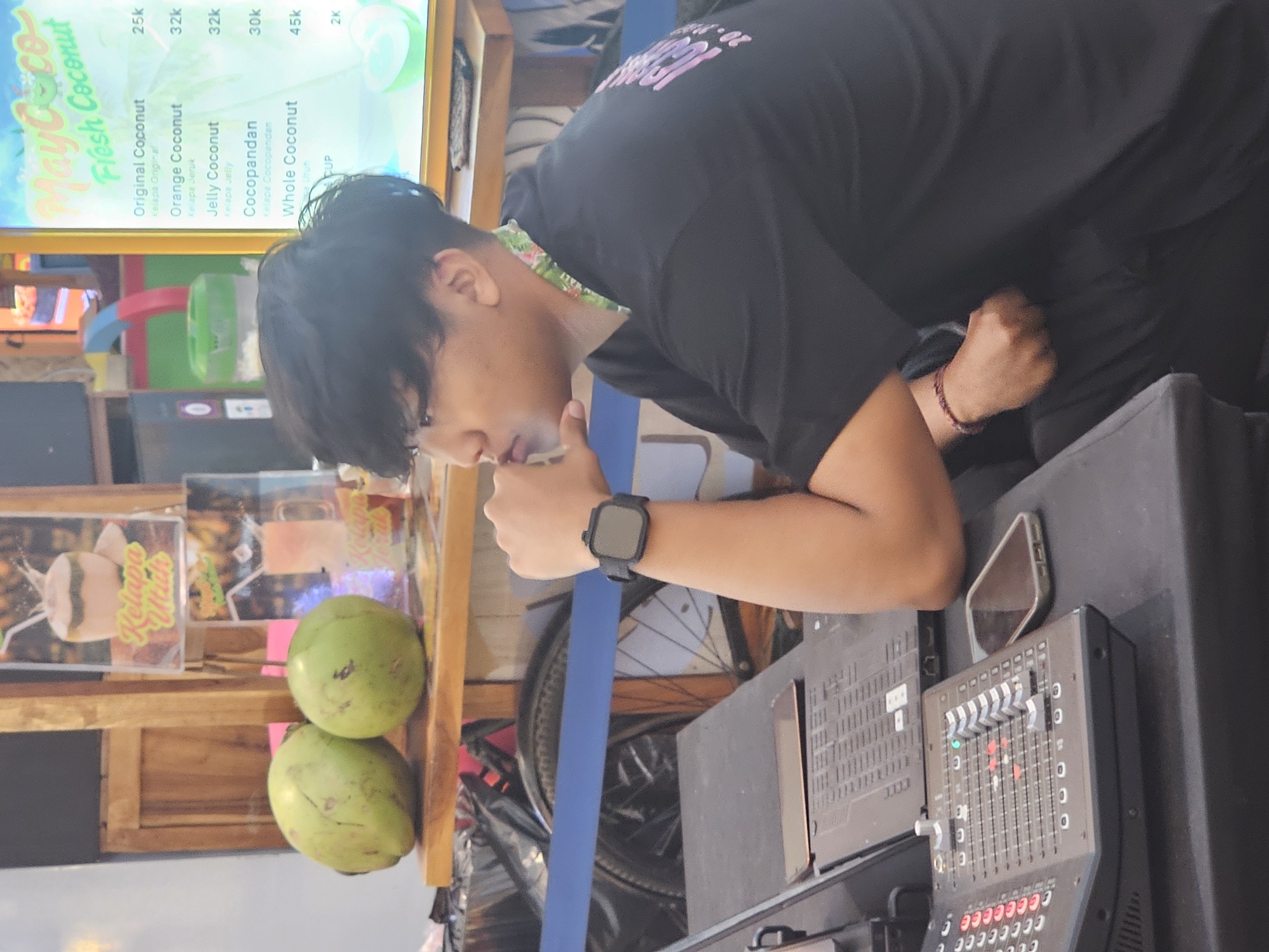 The image shows a man in a black t-shirt leaning over a sound mixing board at a fresh coconut stand in Denpasar, Indonesia. The man is wearing a smartwatch and has his chin resting on his hand. The stand features a menu board listing different coconut drinks with prices: Original Coconut (25k), Orange Coconut (32k), Jelly Coconut (32k), Cocopandan (30k), and Whole Coconut (45k). Two green coconuts are stacked near the stand. A phone is visible next to the sound mixing board. A bicycle wheel is visible in the background, partly obscured by a blue divider.