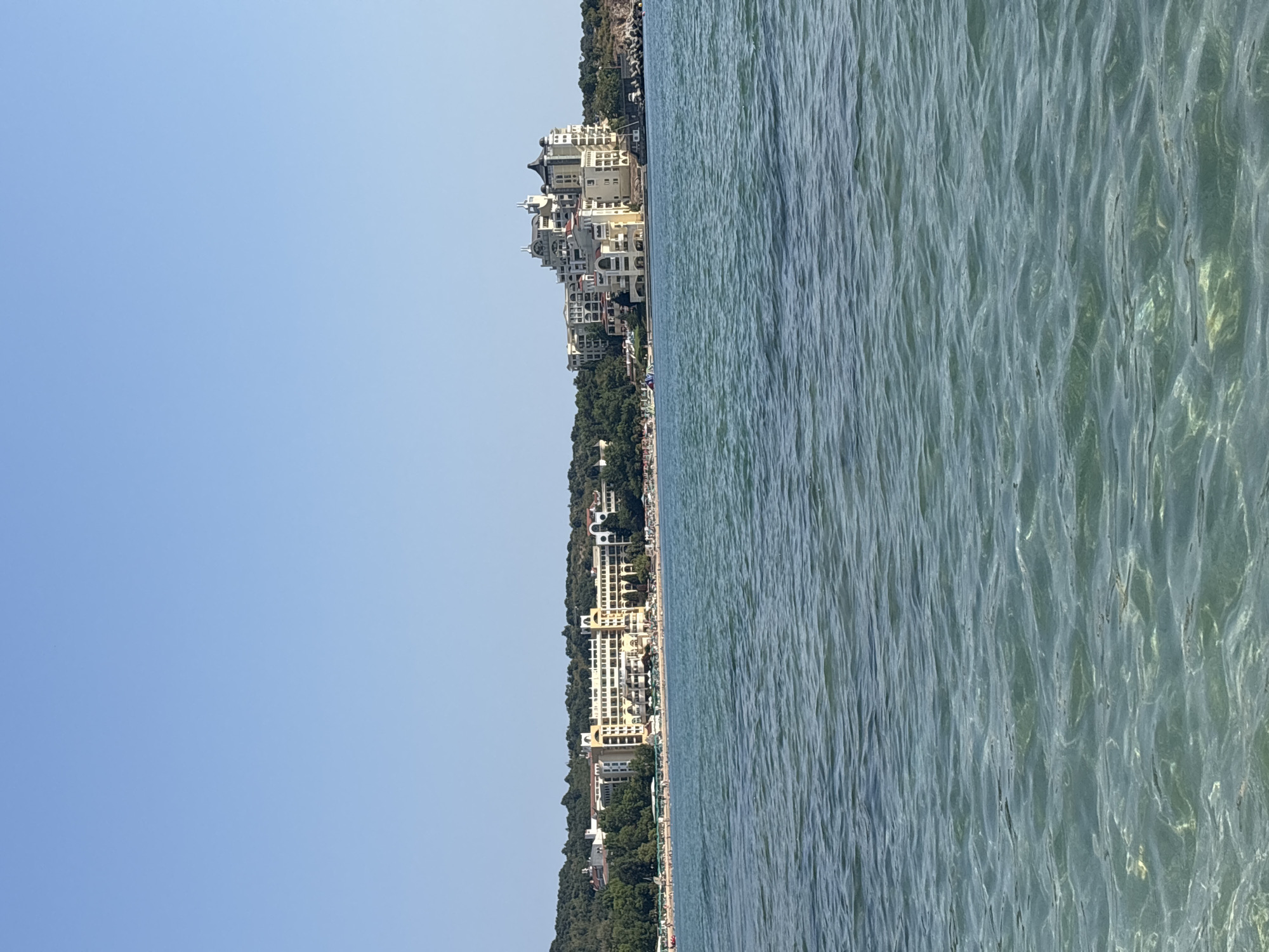 The image shows a panoramic view of the Black Sea coast near Dyuni, Bulgaria. In the foreground, the clear, turquoise water exhibits gentle ripples, with sunlight reflecting off the surface and illuminating the sandy seabed below. In the middle ground, a crowded beach is lined with numerous umbrellas and lounge chairs, indicating a lively summer atmosphere. People are visible scattered across the sand, some walking and others relaxing.
The background features a picturesque coastal landscape dominated by large, multi-story hotels and resort buildings. These structures are predominantly cream-colored with white accents and feature a distinct architectural style with numerous balconies, arches, and varied rooflines. The hotels are nestled against a backdrop of lush green hills covered with dense trees, suggesting a natural environment merging with the developed resort area. The sky above is a clear, bright blue, characteristic of a sunny day. The scene captures a popular tourist destination, highlighting the blend of natural beauty and resort infrastructure.