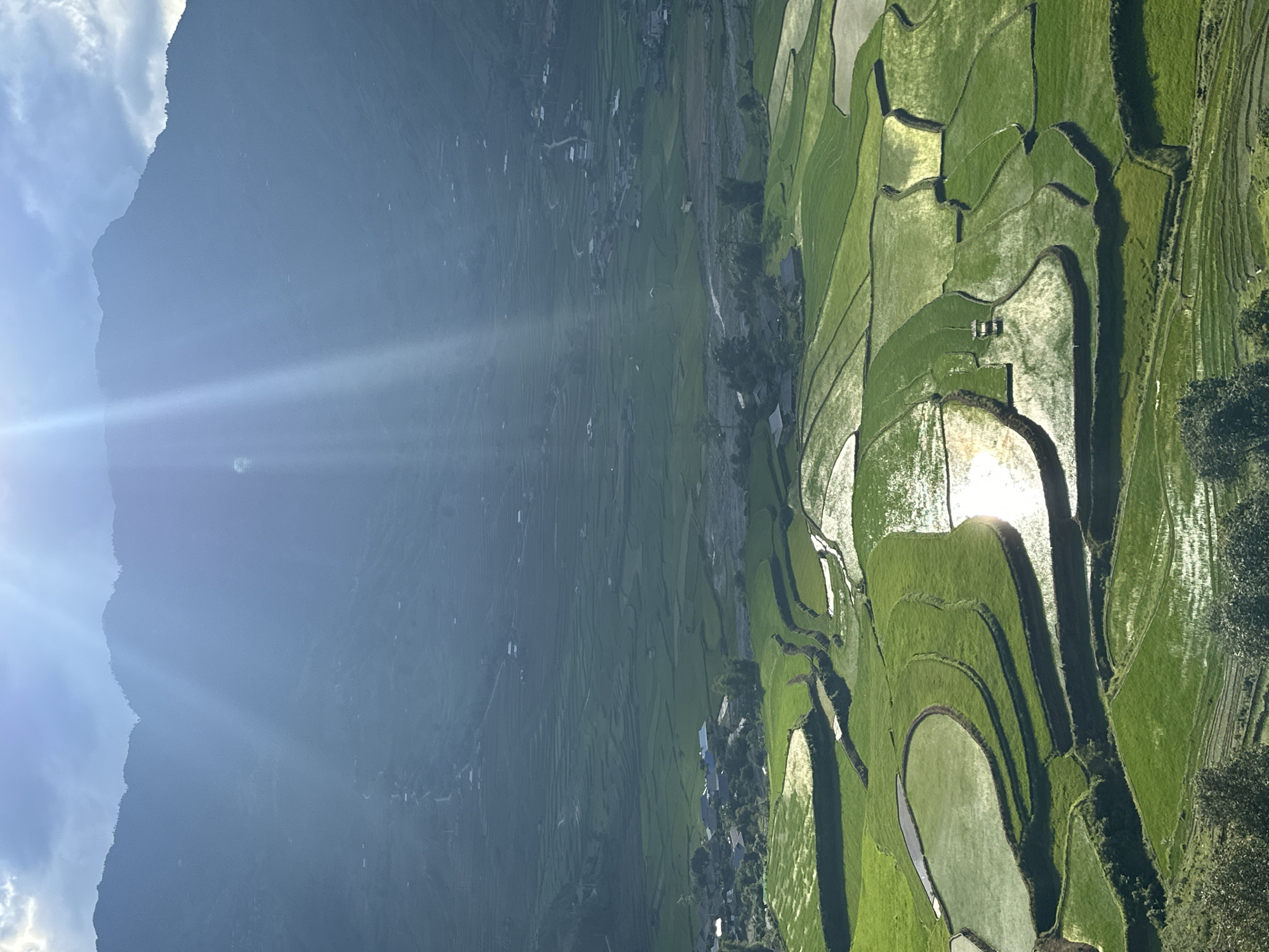 This image captures a panoramic view of the terraced rice fields in the valley of Tú Lệ, Vietnam, under a bright, partly cloudy sky. Intense sunlight from the upper left casts prominent rays and causes significant lens flare. Distant mountains, hazy and blue-grey, form the backdrop, their peaks outlined against the lighter sky.
The middle ground features a sprawling valley floor with numerous terraced rice paddies, some filled with reflective water that mirrors the sunlight, while others are vibrant green with young crops. A winding, light-colored river or stream meanders through the valley, and several clusters of houses with dark roofs indicate small settlements are interspersed among the fields. Unpaved roads or paths connect these settlements and traverse the valley. The immediate foreground shows dark green foliage, suggesting an elevated vantage point. No individuals or specific actions are discernible.