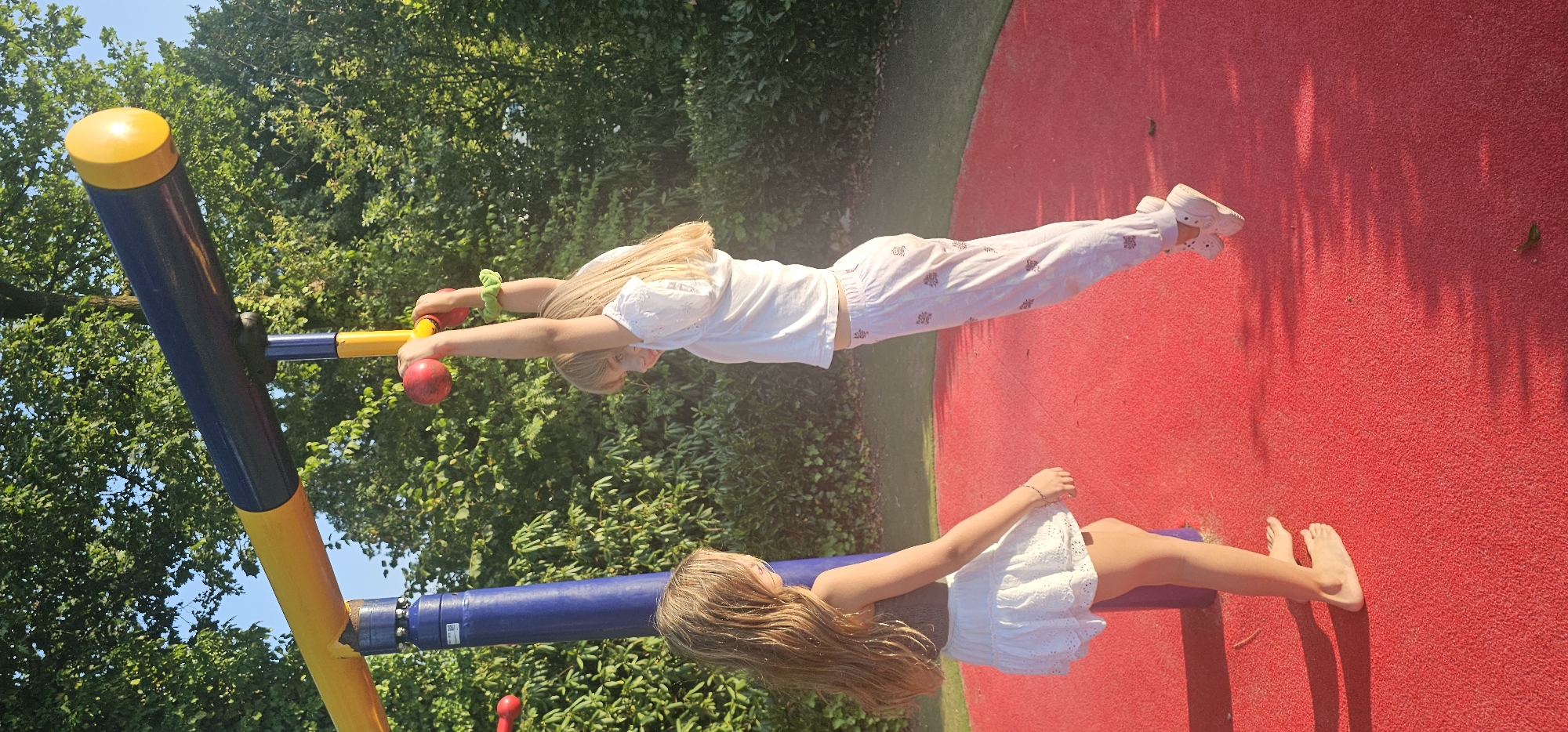 Two young girls are playing on a playground in Groenlo, Netherlands. The girl on the right is hanging from a yellow bar with red handles, her arms outstretched. She is wearing a white shirt and white pants with a subtle pink pattern. The girl on the left is standing and looking up at the other girl. She is wearing a white skirt and a brown top. They are on a red, rubberized surface. The background consists of green trees and a blue sky. The sun is casting shadows on the ground.