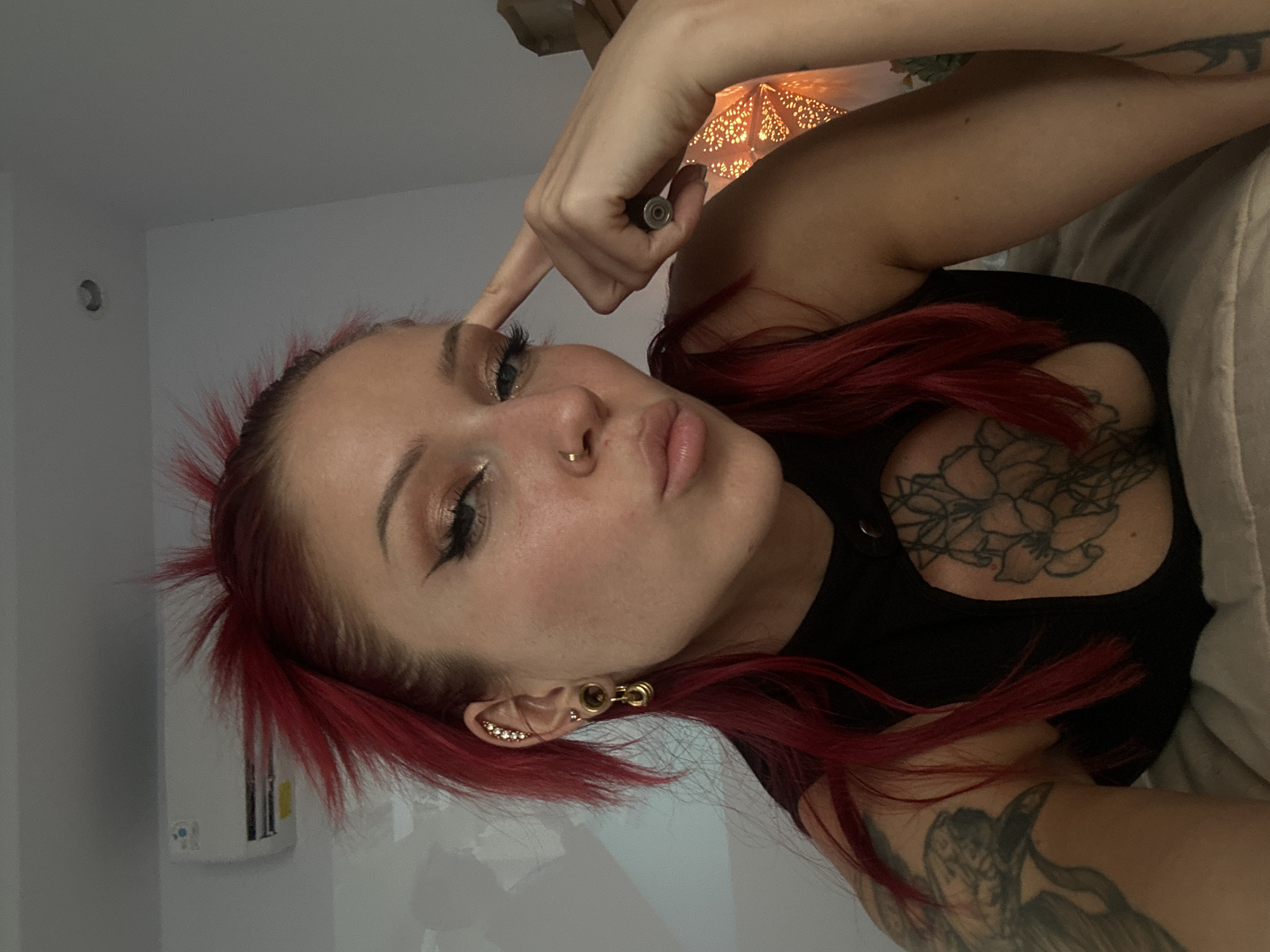 A woman with red hair is looking at the camera. She has fair skin, dark eye makeup, a nose ring, and multiple ear piercings. She's wearing a black top with a cutout at the chest, revealing a floral tattoo. She's holding a small, cylindrical object near her eyebrow with her right hand. Tattoos are visible on her arm. She is lying down on a light-colored bedding with a lit lamp visible in the background. The location is uncertain, with no clear indication of Dalung, Indonesia.