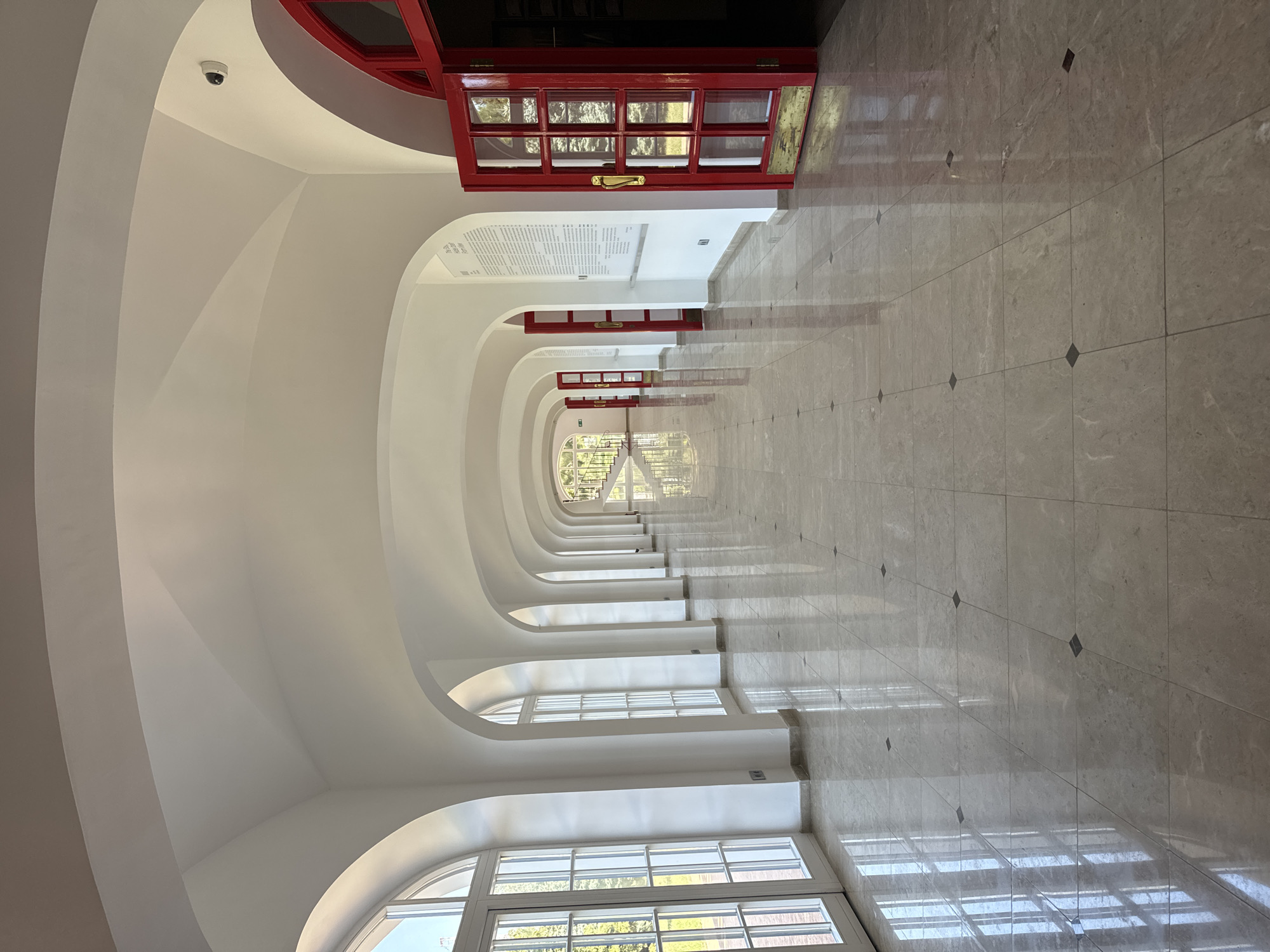 A long, bright white hallway with arched ceilings and multiple arches on the left side with large windows. The floor is made of polished marble tiles with small dark square accents. On the right side, there are multiple red doors, some open, leading into darker rooms. There is a security camera visible on the ceiling. The hallway recedes into the distance where a staircase is visible. The overall impression is one of grand, classic architecture with modern touches.