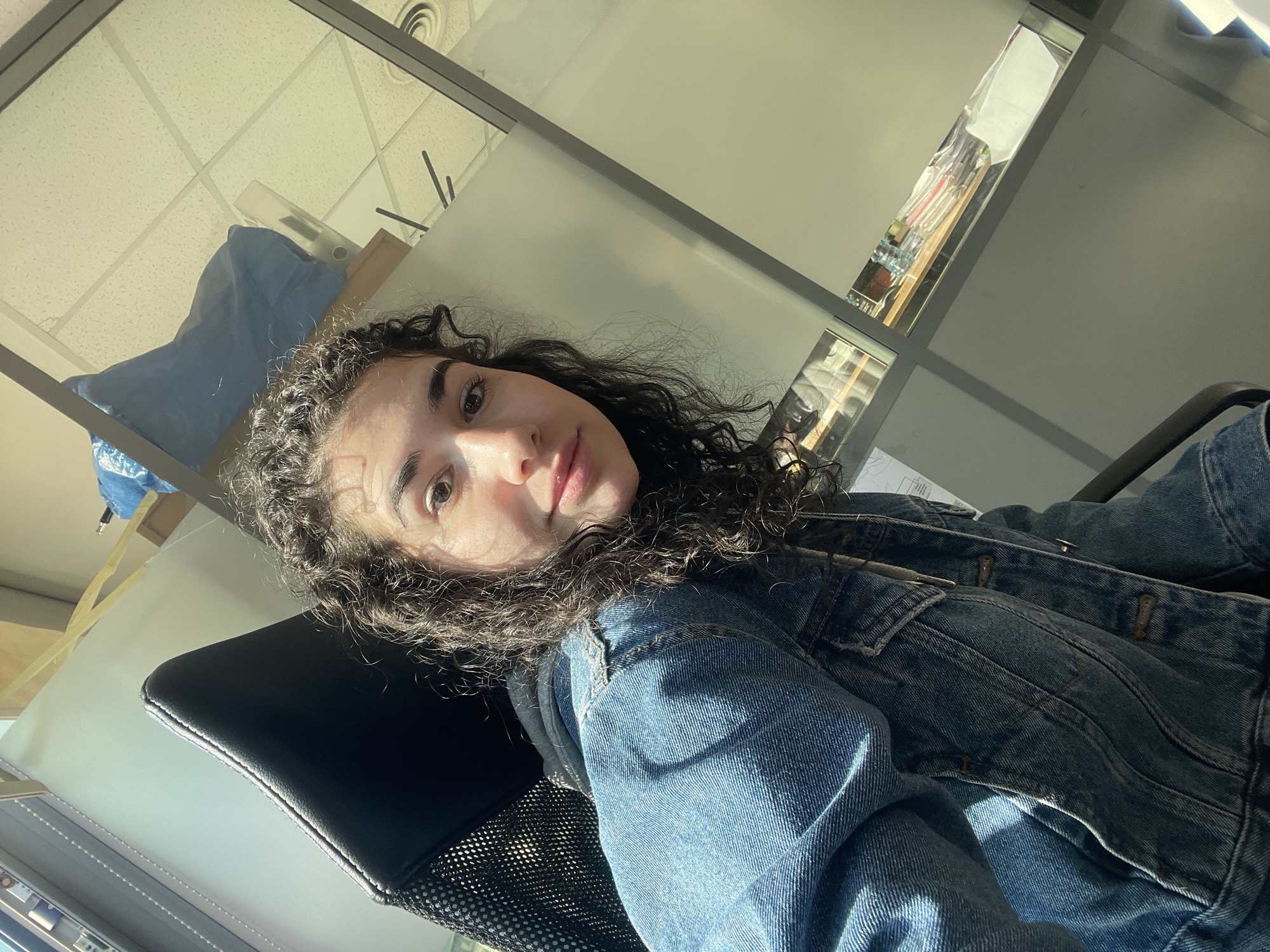 This media file is a low-angle selfie, slightly rotated, featuring a young person with medium-dark, curly hair, likely female. The individual is looking directly at the camera with a neutral, relaxed expression, possibly a slight smirk. They are wearing a blue denim jacket over a dark-colored top, seated in a black office chair with a mesh back.
The setting appears to be an indoor office or commercial space. The background features a light-colored false ceiling with square panels and a circular air vent. There are reflections in glass partitions or windows, revealing abstract glimpses of shelves, papers, and other items within the office. In the upper left background, a blue object, possibly a bag or draped fabric, is visible next to some light-colored structures.
The scene is brightly lit by natural light, likely sunlight, entering from a side angle. This creates strong highlights on the person's face and the denim jacket, and casts noticeable shadows, indicating it is daytime. The activity is a self-portrait or selfie. No discernible text is visible anywhere in the image. While the location context provided is Sofia, Bulgaria, there are no specific visual cues within the image itself to confirm this.