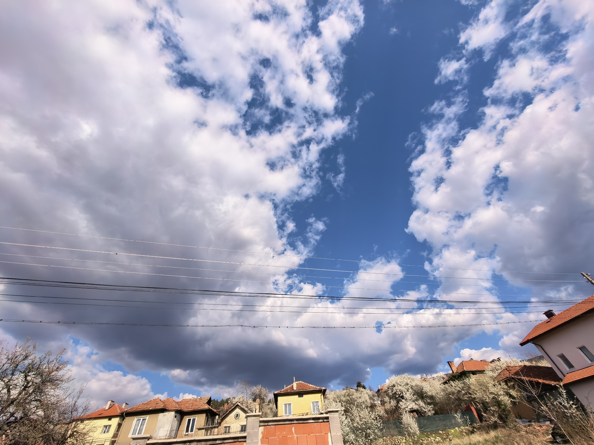 A low-angle shot captures a residential area under a dynamic sky in Unknown, Unknown. Above a row of houses with red-tiled roofs, a mixture of dark and light clouds dominate the upper portion of the frame, with patches of blue sky peeking through. Power lines stretch horizontally across the center of the image, slightly obscuring the view of the buildings below. In the foreground, houses of varying heights and colors, including yellow and beige, are visible, some with unfinished brickwork. Trees with white blossoms dot the landscape, adding a touch of springtime. The scene suggests a partly cloudy day in a quiet neighborhood.
