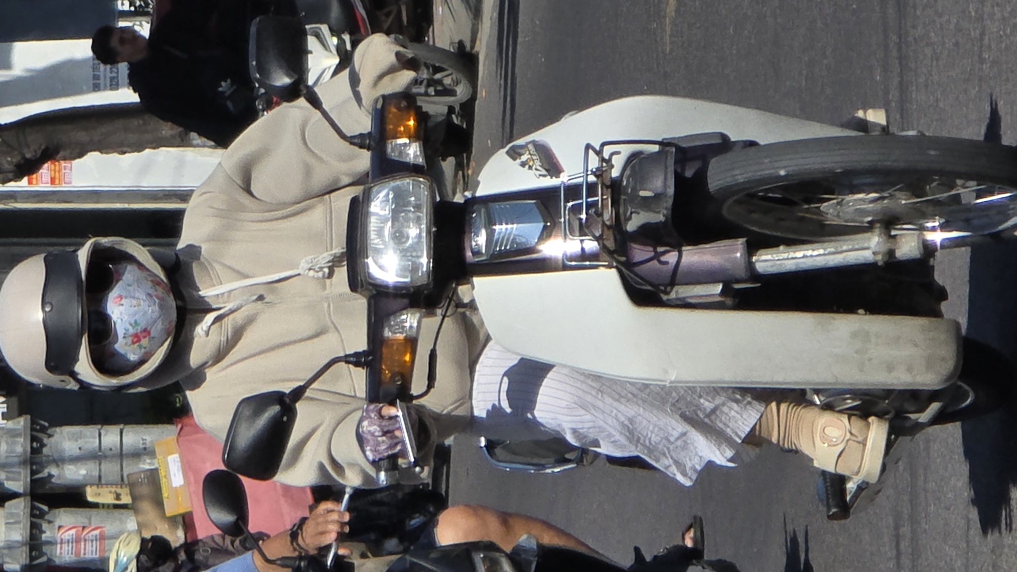 A motorcyclist in Bắc Nha Trang, Vietnam, wears a beige helmet with a dark band and a patterned face mask. The person is also wearing a light beige hoodie, a striped white and gray shirt, and tan gloves. The motorcycle is predominantly white with a rectangular headlight. The street and other vehicles are visible in the background. Another person in a black jacket is seen nearby.