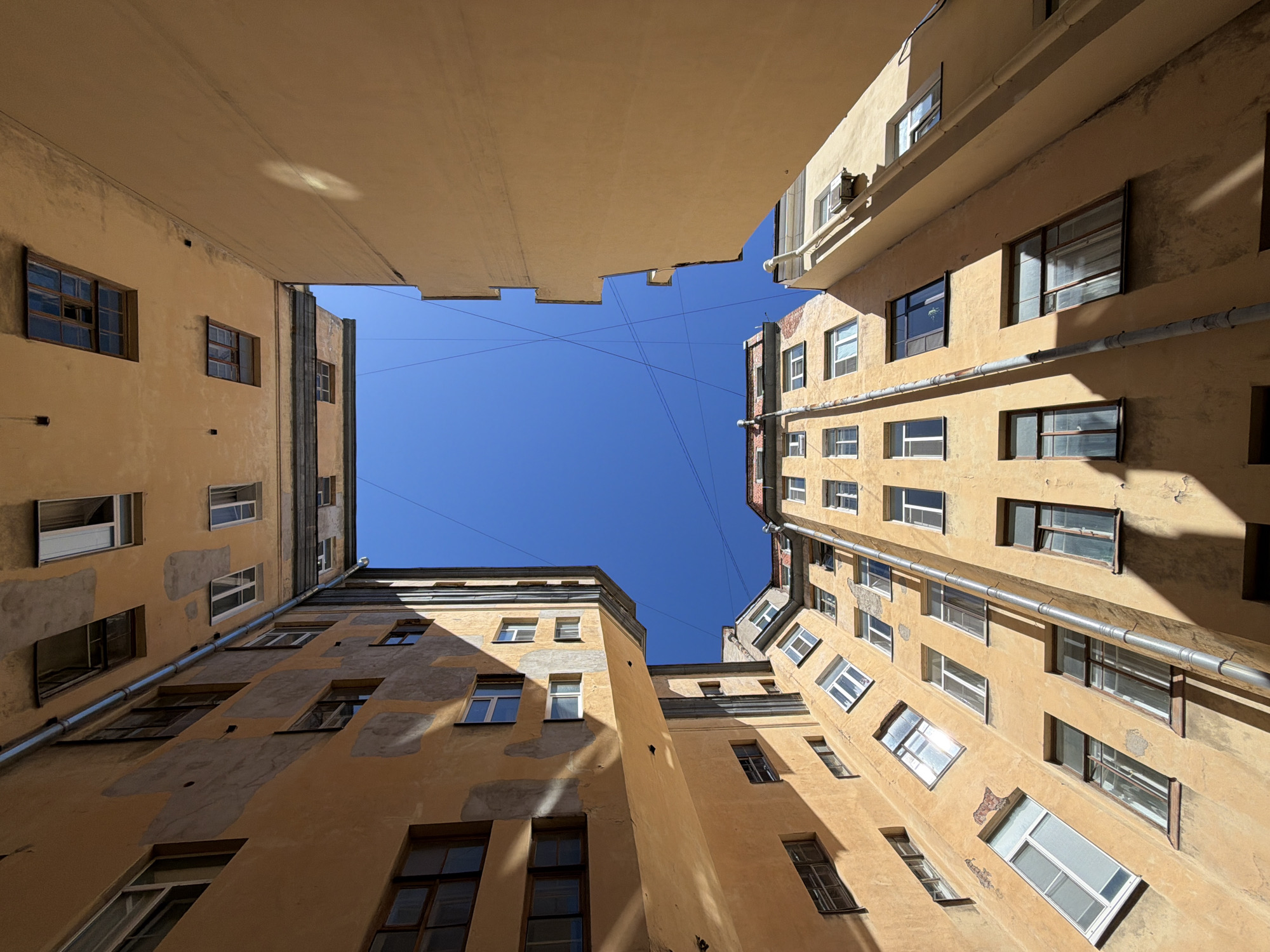 The image presents an extreme upward-angle view into a densely constructed urban courtyard, characteristic of Saint Petersburg, Russia. Multiple multi-story residential buildings frame a central, irregular aperture of clear blue sky.
The building facades are predominantly painted in faded ochre and yellow-orange hues, exhibiting significant signs of age and wear. Large areas of plaster have eroded or peeled away, revealing underlying materials or lighter-colored repair patches. Distinct shadows are cast across various walls, indicating direct and strong sunlight from an unseen source.
Numerous windows are visible on all exposed building surfaces. These windows vary in style, including older wooden-framed, multi-pane units and more contemporary single-pane windows, some of which reflect the bright blue sky. Several vertical, corrugated metal drainpipes are affixed to the building exteriors.
The sky, viewed directly above, is a vibrant, unblemished blue, traversed by two intersecting thin, dark utility wires forming an 'X' pattern. No people, animals, or vehicles are present within the captured scene. The overall impression is one of weathered, densely packed urban architecture under bright daylight.