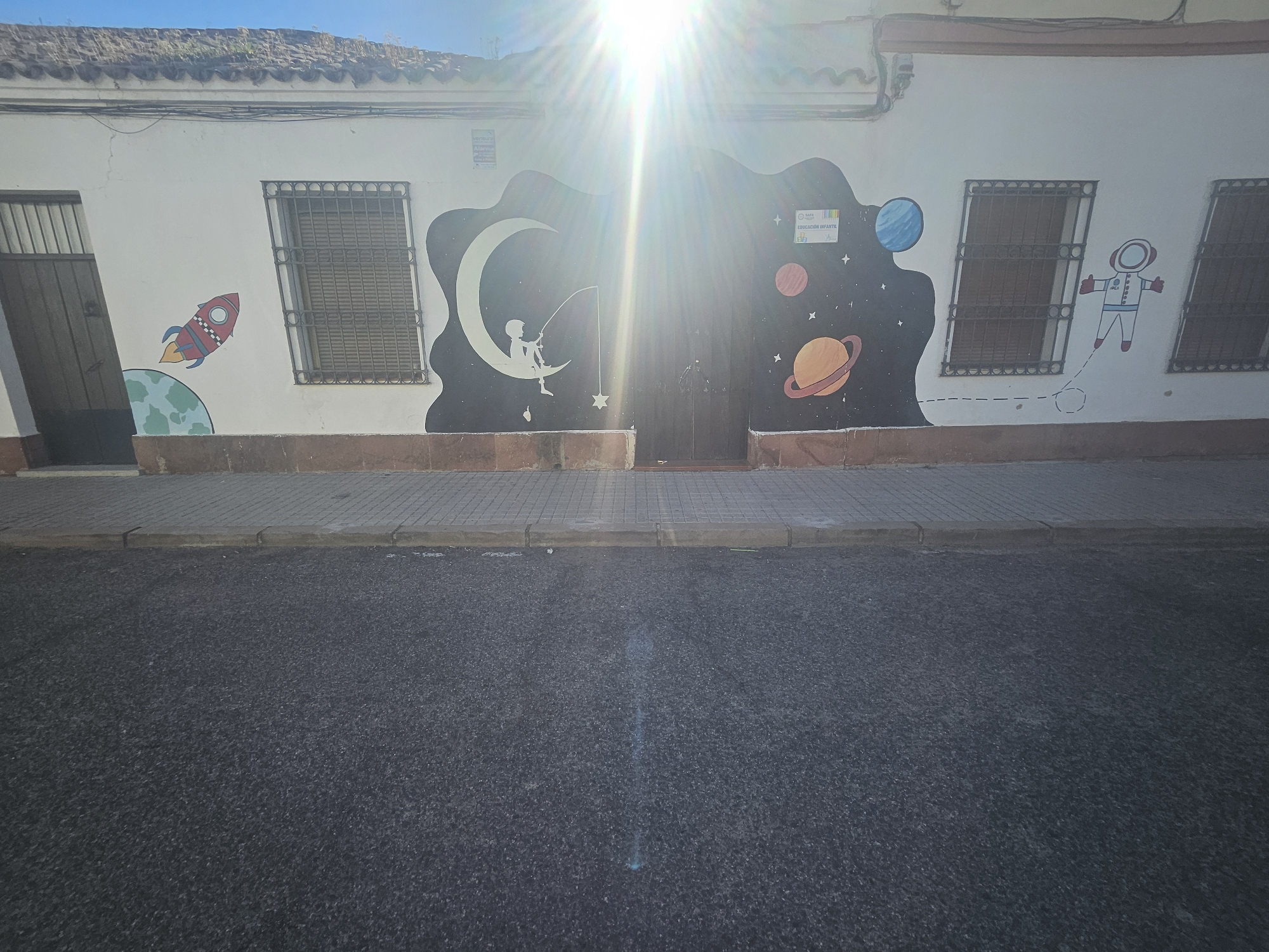 The image shows a street scene in Pedro Abad, Spain. The primary focus is a white building with a mural depicting a space theme. The mural includes images of a rocket ship taking off from Earth, a child fishing from the moon for stars, a blue planet, a red planet, Saturn, and an astronaut floating in space. There are windows with barred security grates on either side of the mural. A door is present in the middle of the mural. The street in front of the building is paved with asphalt and has a tiled curb. The sun shines brightly on the building.