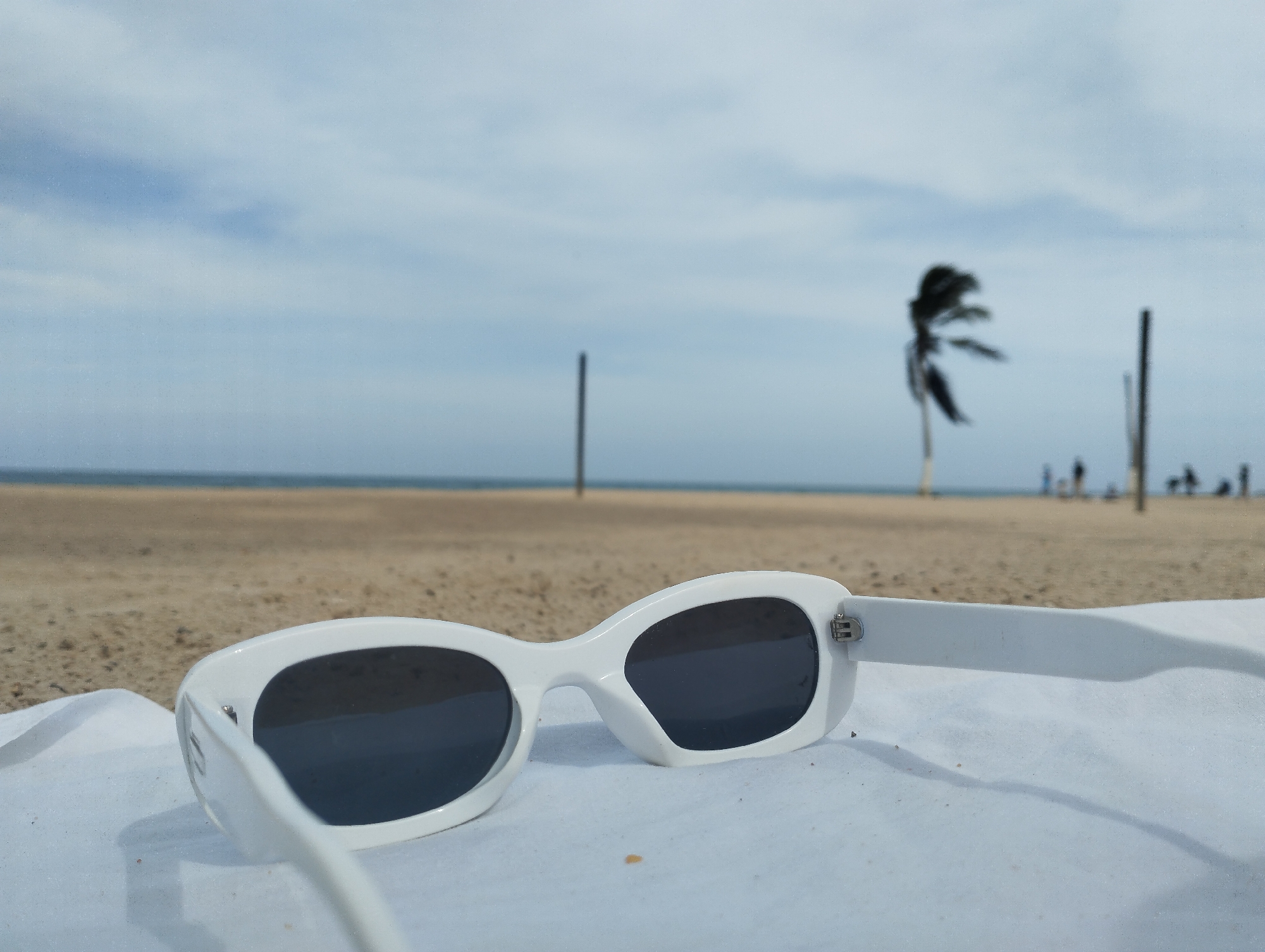 A close-up shot depicts a pair of white-framed sunglasses resting on a white surface, likely a towel, with a beach scene in the background. The beach features light brown sand, and the ocean is visible in the distance under a cloudy sky. A palm tree is prominent on the beach, bending from the wind, with volleyball nets and several indistinct figures in the background. The location is most likely Playa El Agua in Porlamar, Venezuela.