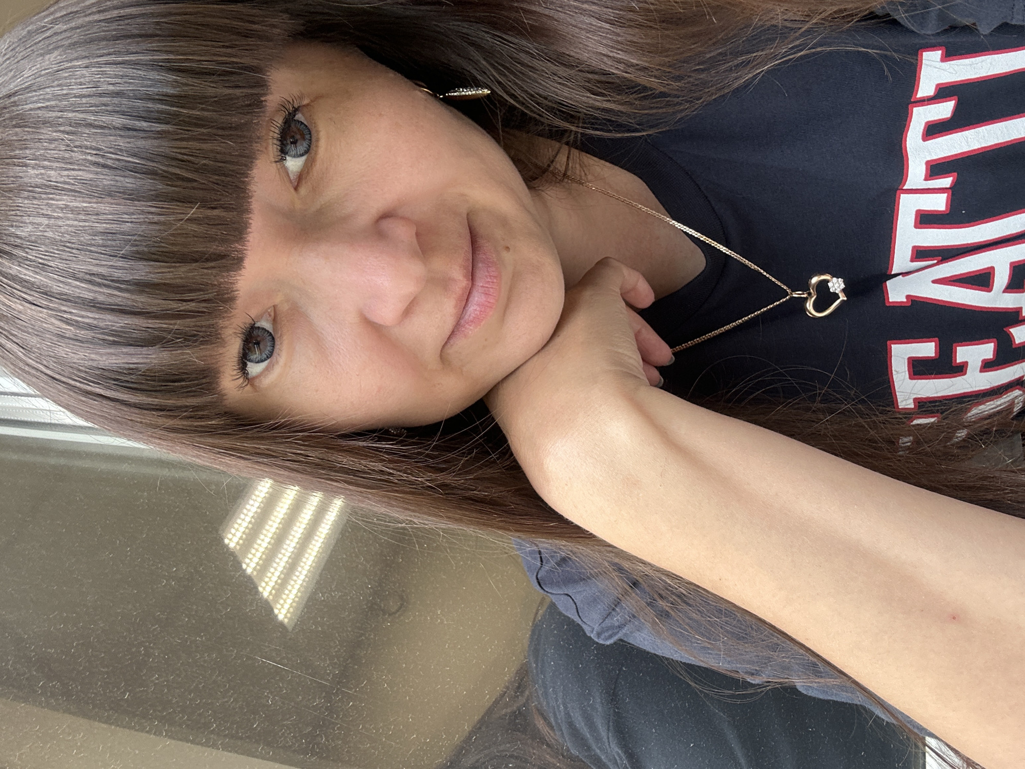 A close-up, head-and-shoulders shot shows a woman with long, dark brown hair and bangs. She is wearing a black t-shirt with red and white lettering that appears to spell out "SEATTLE." She has blue contact lenses and is wearing a gold necklace with a heart-shaped pendant. Her left arm is resting on her chin, and her left hand is supporting her jaw. The background appears to be a window with a reflection of fluorescent lighting. The image was taken in Chaikovsky, Russia.
