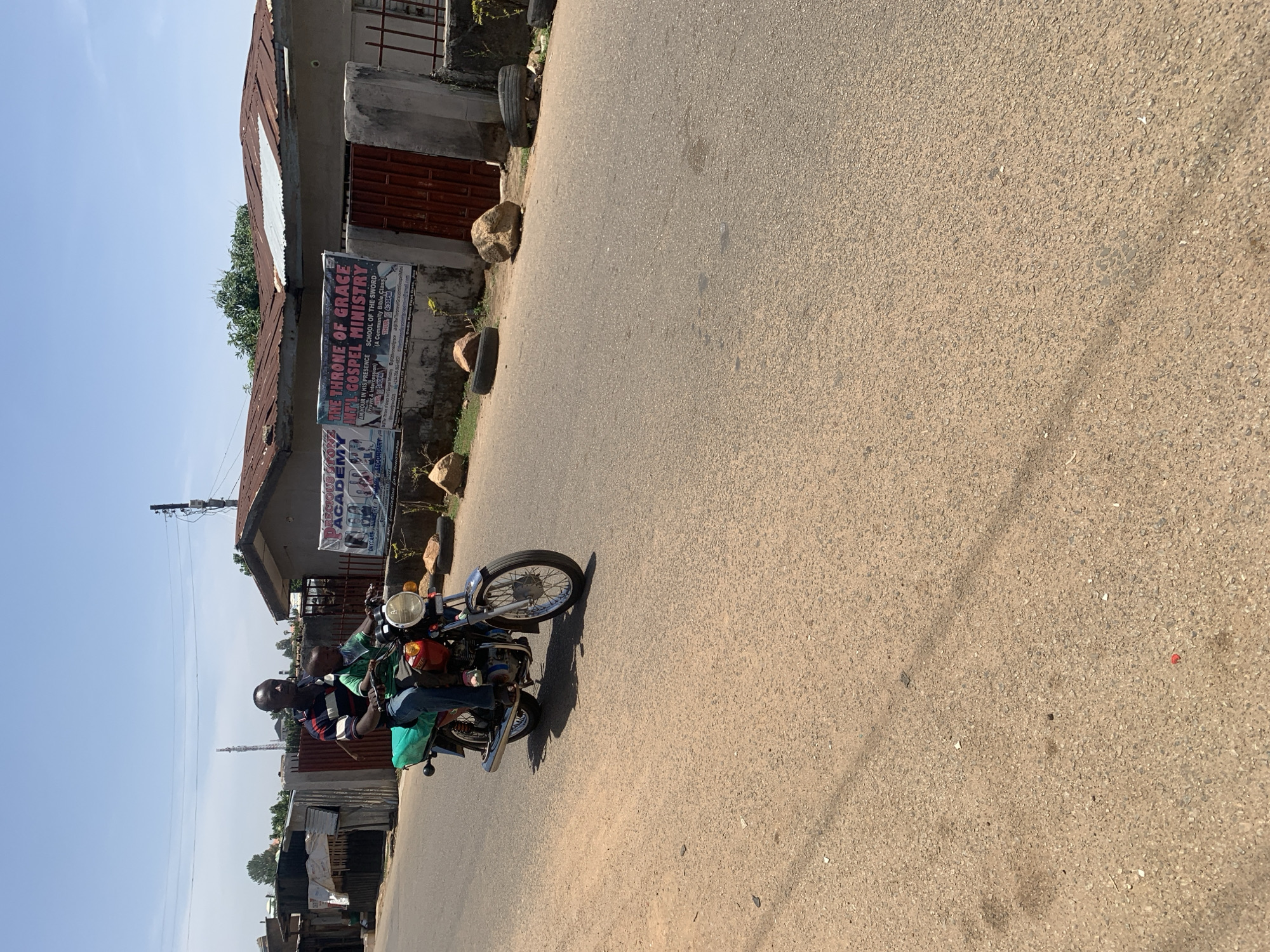 The image depicts a bright, sunny street scene in Jos, Nigeria, featuring two individuals riding a motorcycle on a paved road. The light blue sky is clear, and strong shadows suggest mid-day.
The central subjects are two people, likely men, on a red and black motorcycle. The driver wears a striped shirt and dark trousers, while the passenger behind him is in a green shirt. They are traveling on a dark, coarse-textured asphalt road that shows some wear.
Along the left side of the road, several buildings are visible. The most prominent structure has a concrete base and a corrugated iron roof. Two large banners are affixed to this building. One banner reads "THE THRONE OF GRACE INTAL GOSPEL MINISTRY," advertising "AN HOUR IN HIS PRESENCE (Prayer & Intercession)" and "SCHOOL OF THE SWORD (A Community Bible Class)" with a mention of "THUR 4:30PM." Below it, another banner announces "PRECIOUS STONE ACADEMY," offering "DAYCARE...PRIMARY...SECONDARY" education.
The immediate roadside features dirt, scattered rocks, and a couple of old tires. Further down the street, utility poles and additional simple structures with corrugated roofs can be seen, indicating a populated, semi-urban environment.