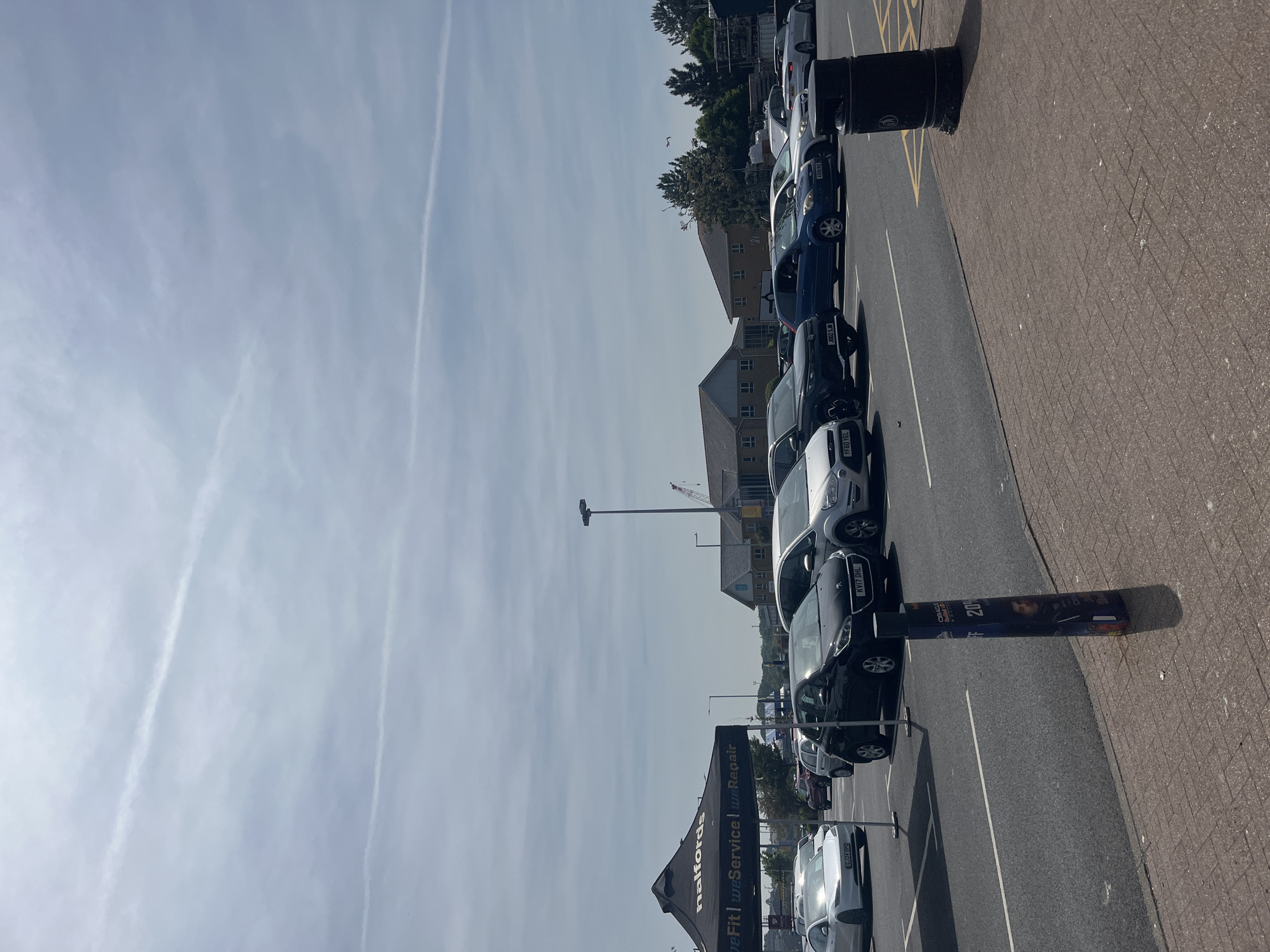 The image depicts a parking lot in East Suffolk, United Kingdom, densely occupied by several cars. The location appears to be adjacent to a building, possibly a retail or commercial establishment. A tall light pole can be seen in the background, along with buildings. A sign for "Halfords," an automotive and leisure retailer, is partially visible in the foreground, indicating that the parking lot is likely associated with the store. Yellow lines and painted parking spaces are seen on the asphalt. The sky above is clear with some clouds. Visible textual details include vehicle license plates (e.g., CJ24 OPD, KV17 DHL, YF60 YEL, J002 SJM) and the words "Service" and "Repair" on the Halfords sign. Also seen is a banner advertising a sports event.