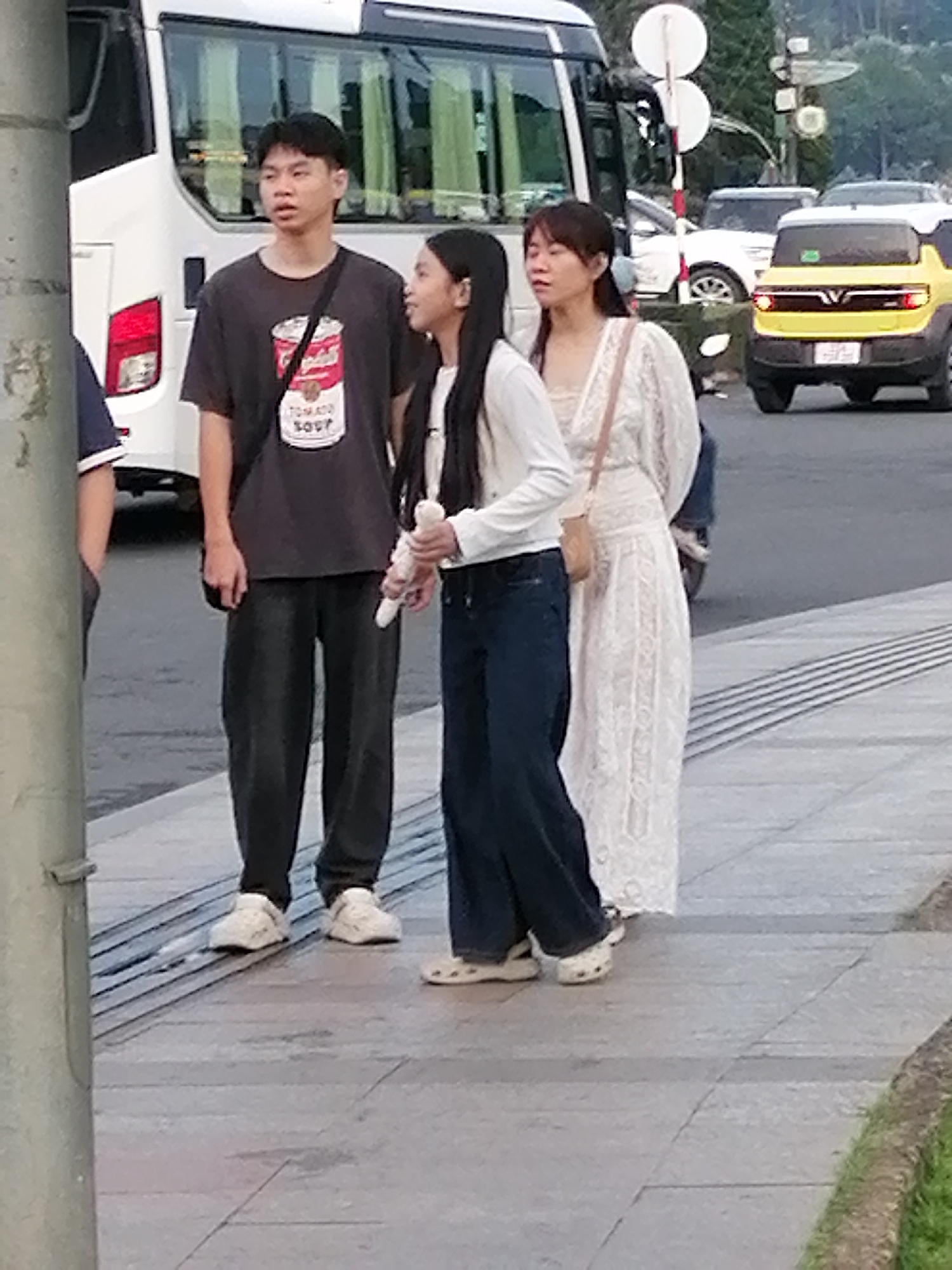 Three individuals are standing on a paved sidewalk in Da Lat, Vietnam, with a white tour bus in the background. A young man on the left, wearing a dark t-shirt with a "Tomato Soup" graphic and dark pants, stands facing left with his mouth slightly open. Beside him, a young girl in a white long-sleeved shirt and dark jeans holds a white object in her right hand and looks forward. To the right of the girl, a woman in a white, lace-detailed dress and dark jeans stands facing forward, with a small light-brown purse slung across her body. The sidewalk has a textured pattern of parallel lines. In the background, cars are visible on a road, including a yellow SUV. Trees and foliage are present in the distance.