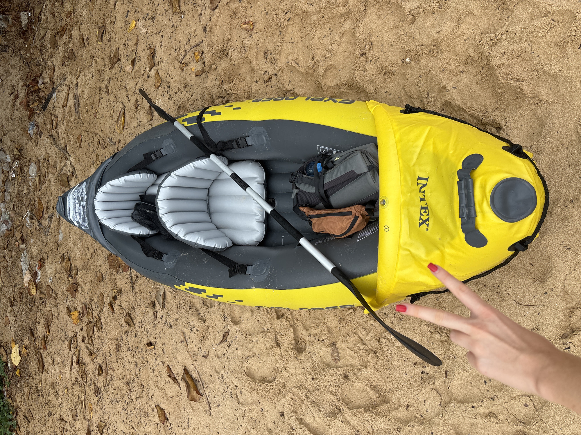 A top-down shot depicts an Intex Explorer K2 kayak on sand, possibly beach sand, with scattered leaves and debris. The kayak is yellow and gray, containing two inflated seats, a two-piece paddle, and two bags. A hand with red nail polish gives the peace sign. The location is likely in Choeng Thale, Thailand.