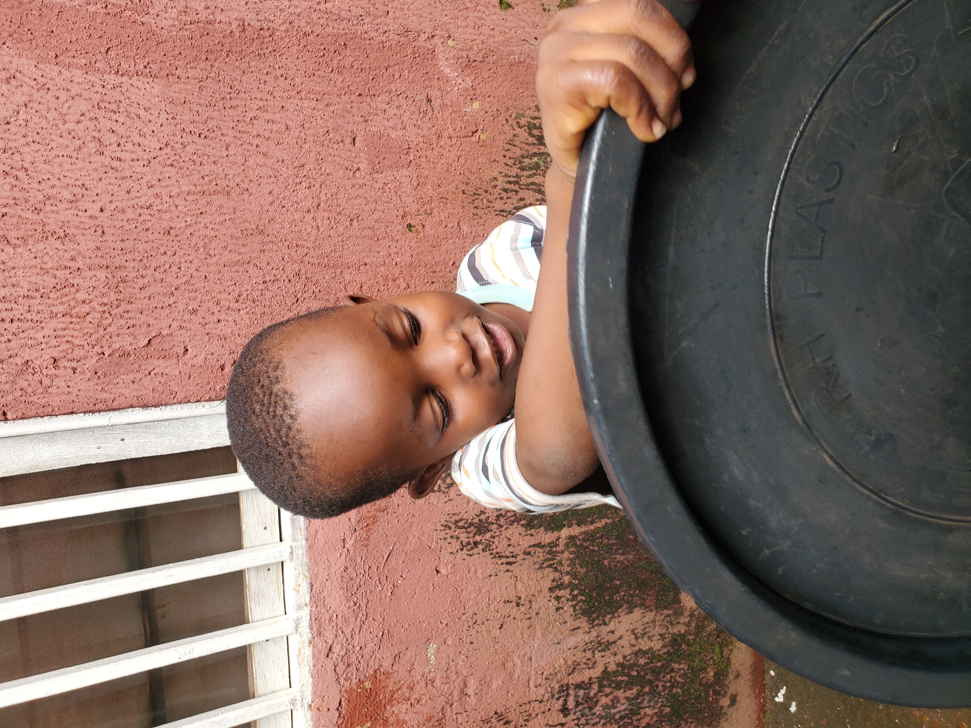 A young boy with dark skin and short, wavy hair is leaning over a large, black plastic basin. He is wearing a horizontally striped shirt with white, yellow, and dark blue bands. His eyes are downcast, and he appears to be looking at something inside or at the edge of the basin. His right hand is gripping the rim of the basin. The background consists of a textured, reddish-brown wall and a section of a window with white bars. There is a hint of greenery or moss on the lower part of the wall. The image appears to be taken outdoors, possibly in a residential area in Jos, Nigeria.