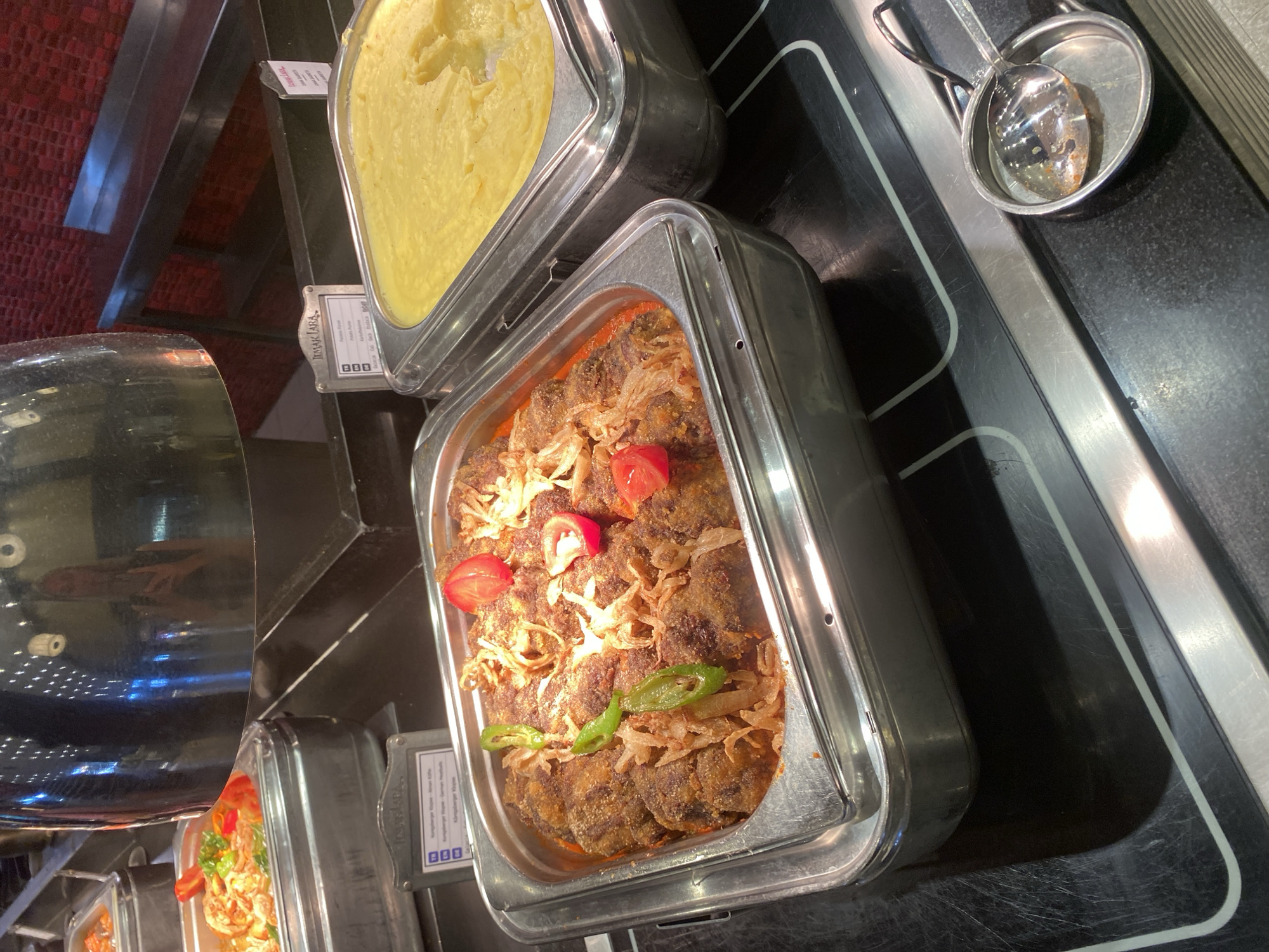The image shows a buffet line in Aksu, Turkey. In the foreground, a metal chafing dish contains brown meatballs in a reddish sauce, topped with fried onions, sliced green peppers, and tomato wedges. To the right of this dish is another metal chafing dish filled with yellow mashed potatoes. A serving spoon rests in a small metal container beside the mashed potatoes. In the background, another chafing dish with food is partially visible, as is a large, reflective black dome over a heating element. A textured red wall and a black countertop are also visible in the background. A person's reflection can be seen in the black dome. A label with text in Turkish, English, and German is visible in front of the meatball dish.