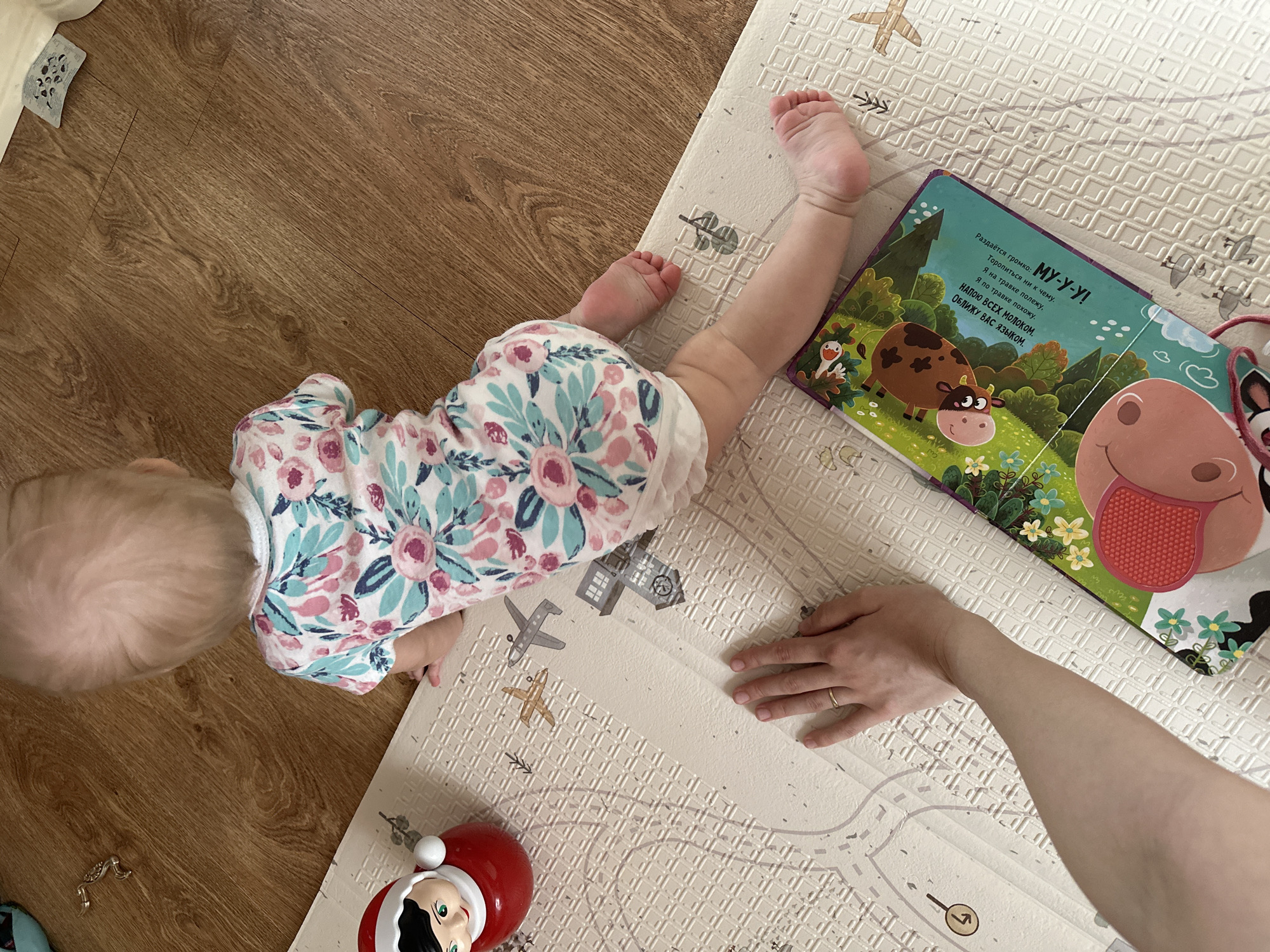 A baby wearing a white floral print onesie is lying prone on a light-colored play mat with road and vehicle designs. The baby's head is turned away from the viewer, showing the back of its head. Next to the baby is an open children's book with a cow illustration and Russian text: "Раздаётся громко: МУ-У-У! Торопиться ни к чему. Я на травке полежу, Я по травке похожу. НАПОЮ ВСЕХ МОЛОКОМ. ОБЛИЖУ ВАС ЯЗЫком." A fair-skinned adult hand is also on the mat near the book. A red toy resembling an elf is on the mat. The mat partially covers a wooden floor.