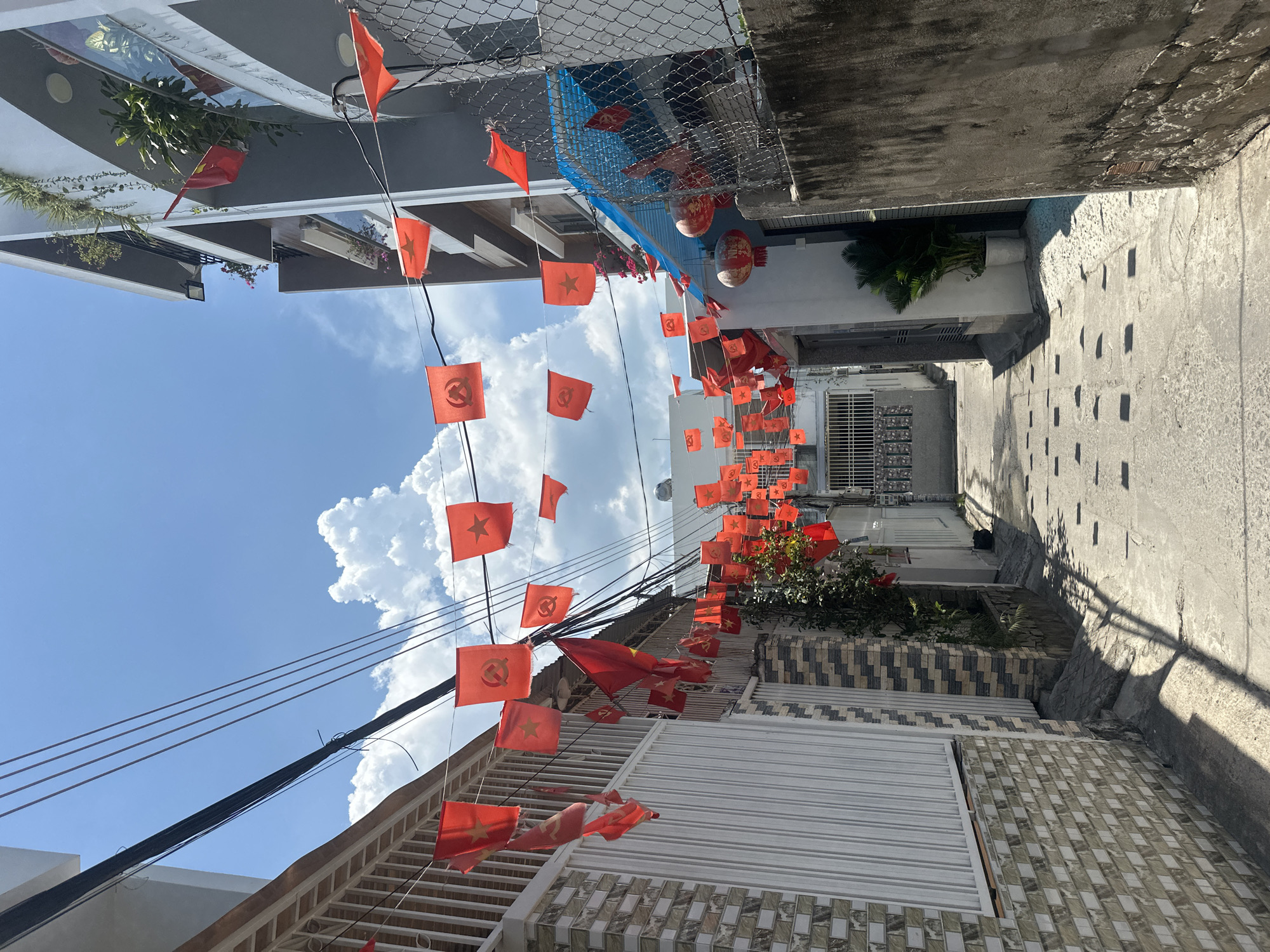 The image shows a narrow alleyway in Bac Nha Trang Ward, Vietnam. Red flags, some with a star and others with a hammer and sickle, are strung across the alleyway. Buildings line both sides of the alley, with a mix of white, gray, and tiled facades. Overhead, power lines are visible against a blue sky with puffy white clouds. The alley floor is paved with concrete. The perspective is looking straight down the alley, creating a vanishing point effect.