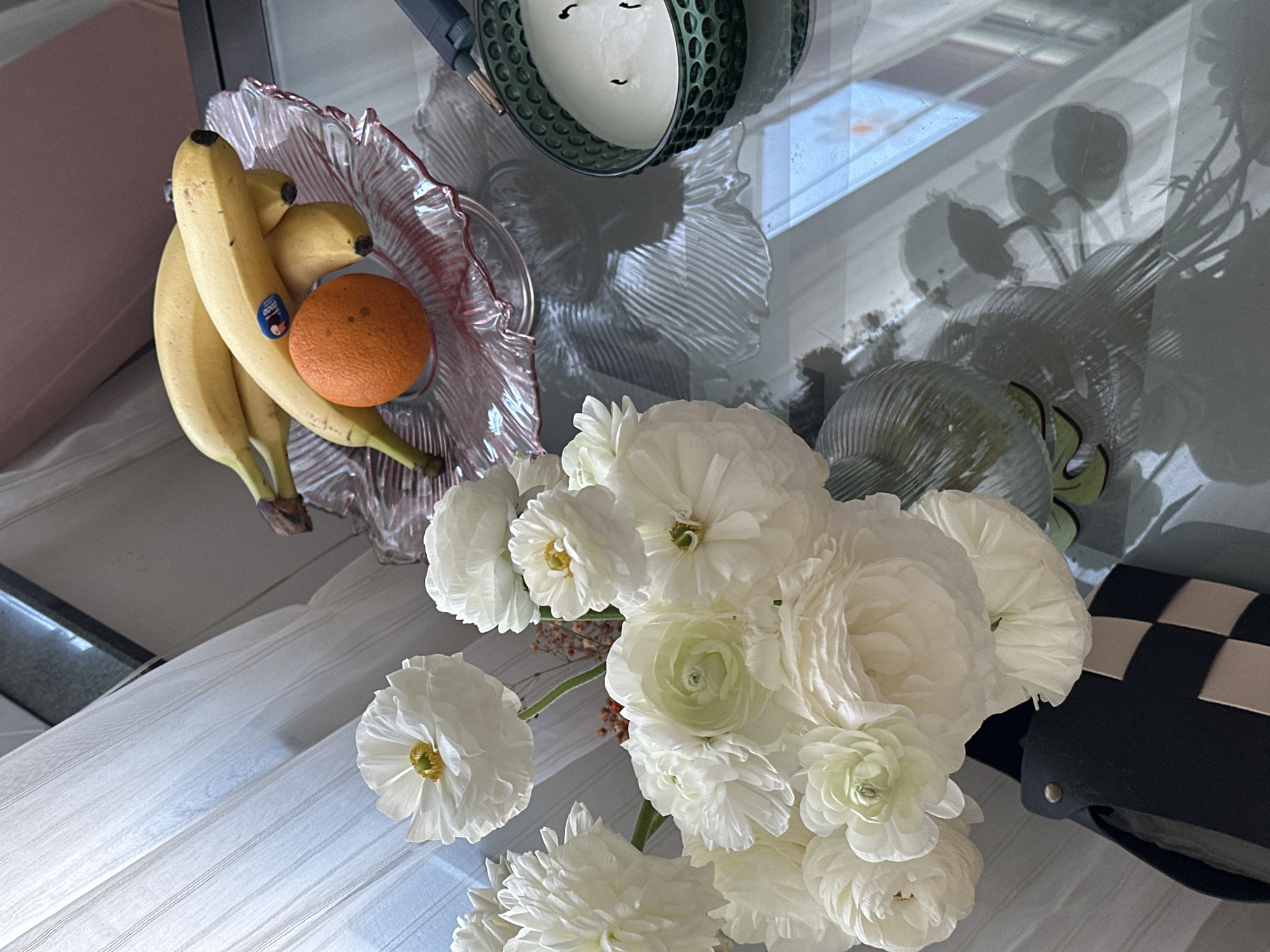 A high-angle shot displays an arrangement of objects on a glass table. A fruit bowl holds bananas and an orange. Adjacent is a green candle holder and a vase containing a bouquet of white ranunculus flowers. A black and white checkered bag is positioned near the flowers. The scene is situated in Novi Sad, Serbia. The environment consists of a table surface with reflections of the external surroundings.