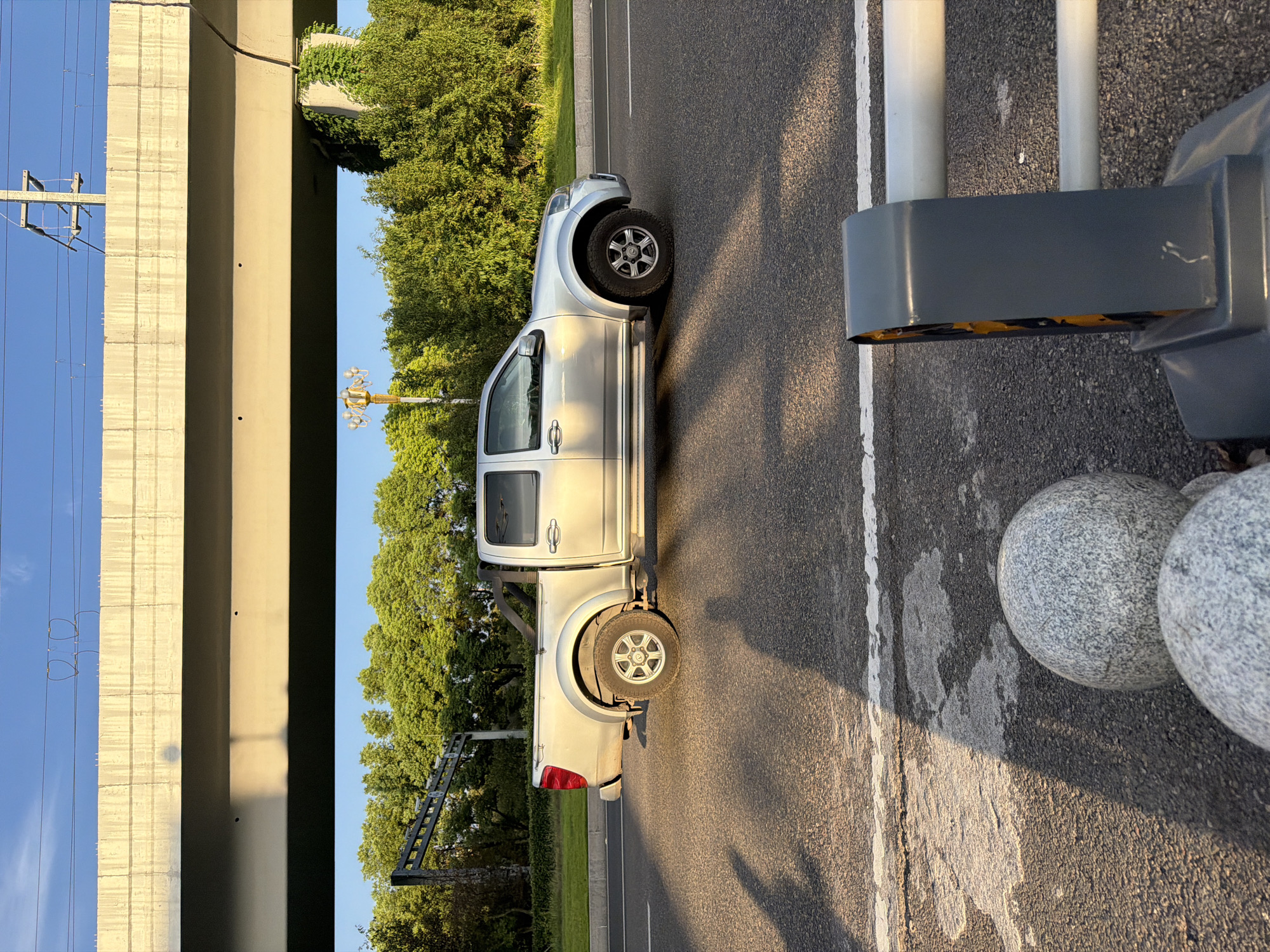 In the city of Jiangjin, China, a silver pickup truck is captured in profile, driving on a road with a clear blue sky overhead. The truck is positioned on the right side of the frame, with its front facing towards the right. The road is paved with asphalt and features white lane markings. A concrete overpass stretches across the upper portion of the image, casting a shadow on the road and truck. Green trees line the side of the road behind the truck, with some vegetation also visible on a pillar supporting the overpass. In the foreground, out of focus, are two spherical granite bollards and a grey traffic barrier with yellow reflective elements. The scene appears to be illuminated by the warm light of late afternoon or early morning sun, creating long shadows across the road. There are no people clearly visible in the image, although shadows on the road suggest the presence of pedestrians or other vehicles not fully within the frame.