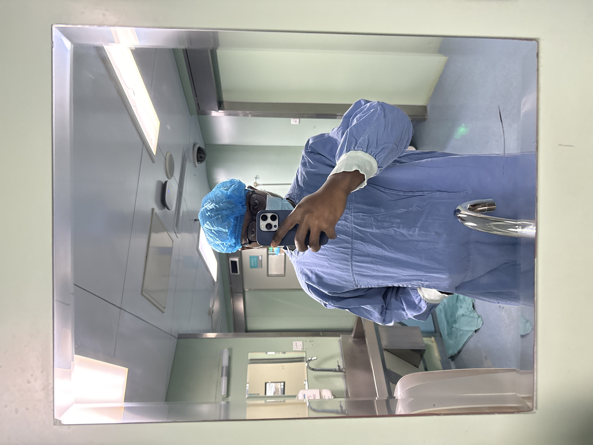 A person in surgical scrubs, including a surgical cap and mask, is taking a selfie in a mirror. The reflection shows a surgical room or pre-op area with stainless steel surfaces and equipment. The precise location in the city of Yuzhong District, China is not ascertainable from this image.
